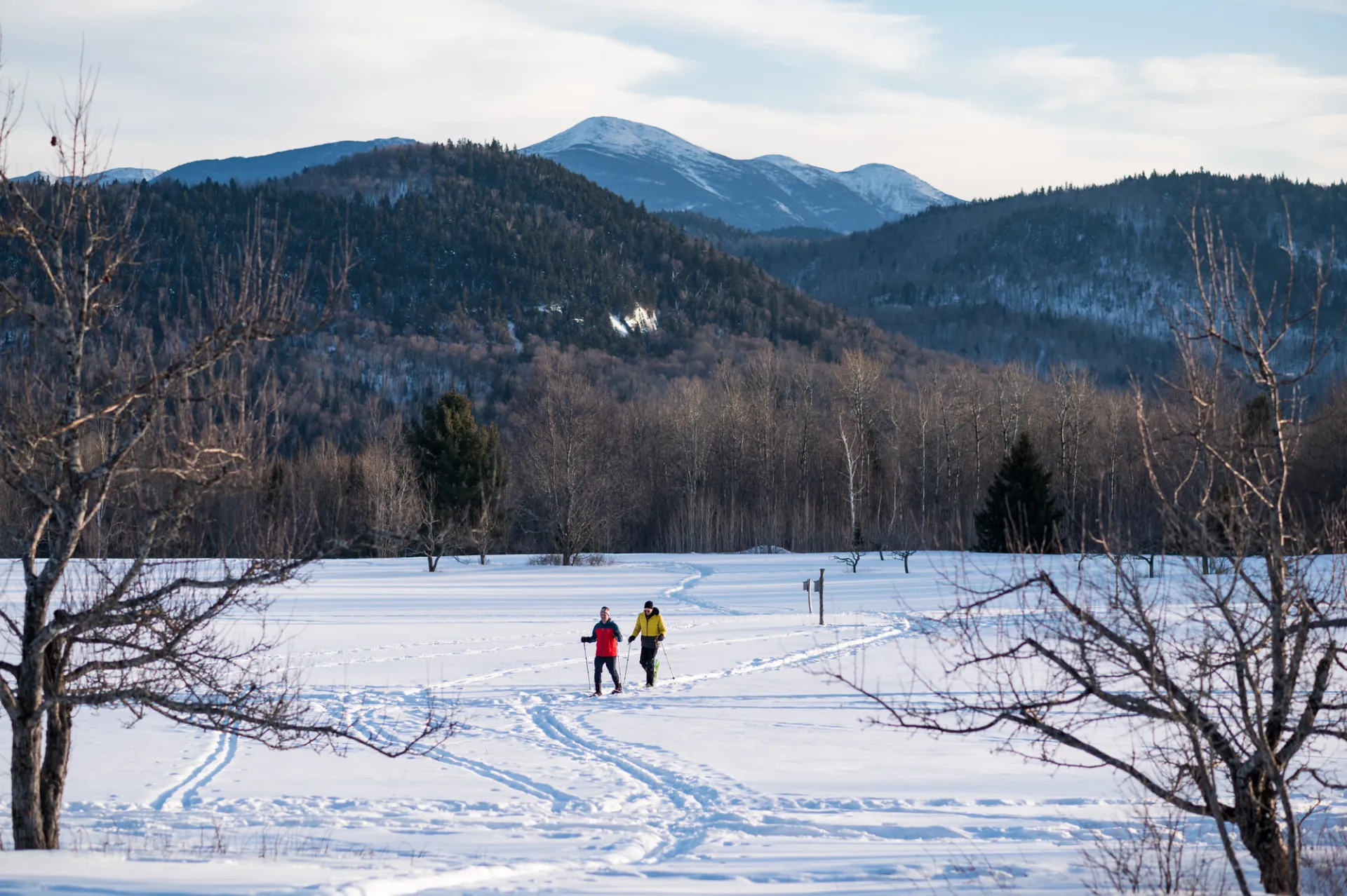 Two people cross-country skiing through a snowy field with mountain views behind them.
