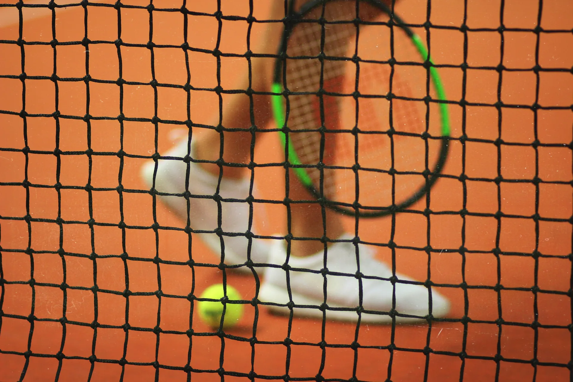 Person's feet in white tennis shoes, racket, and tennis ball seen on clay court through tennis net