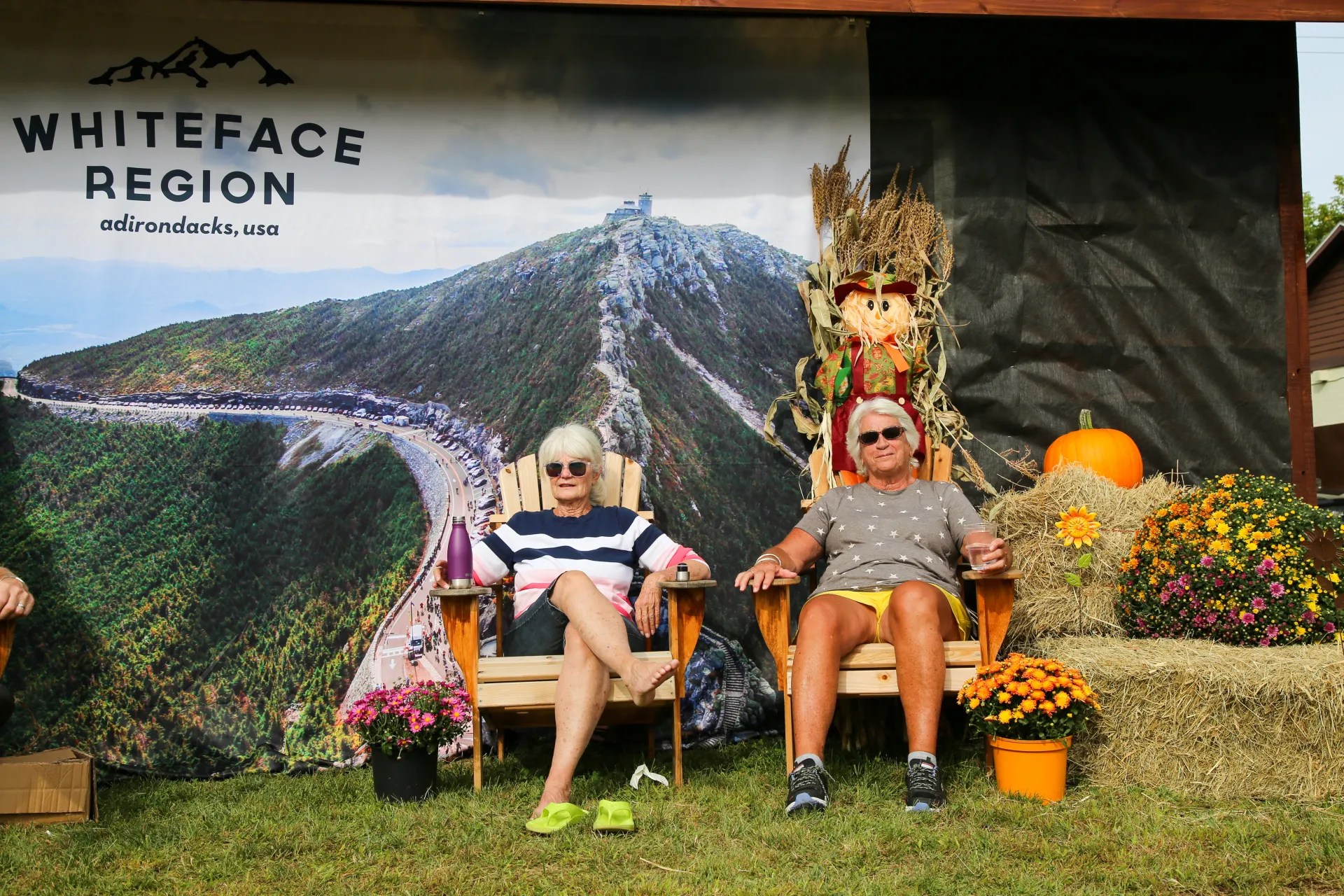 Two women sit in wooden Adirondack chairs surrounded by pumpkins, mums, and hay bales.