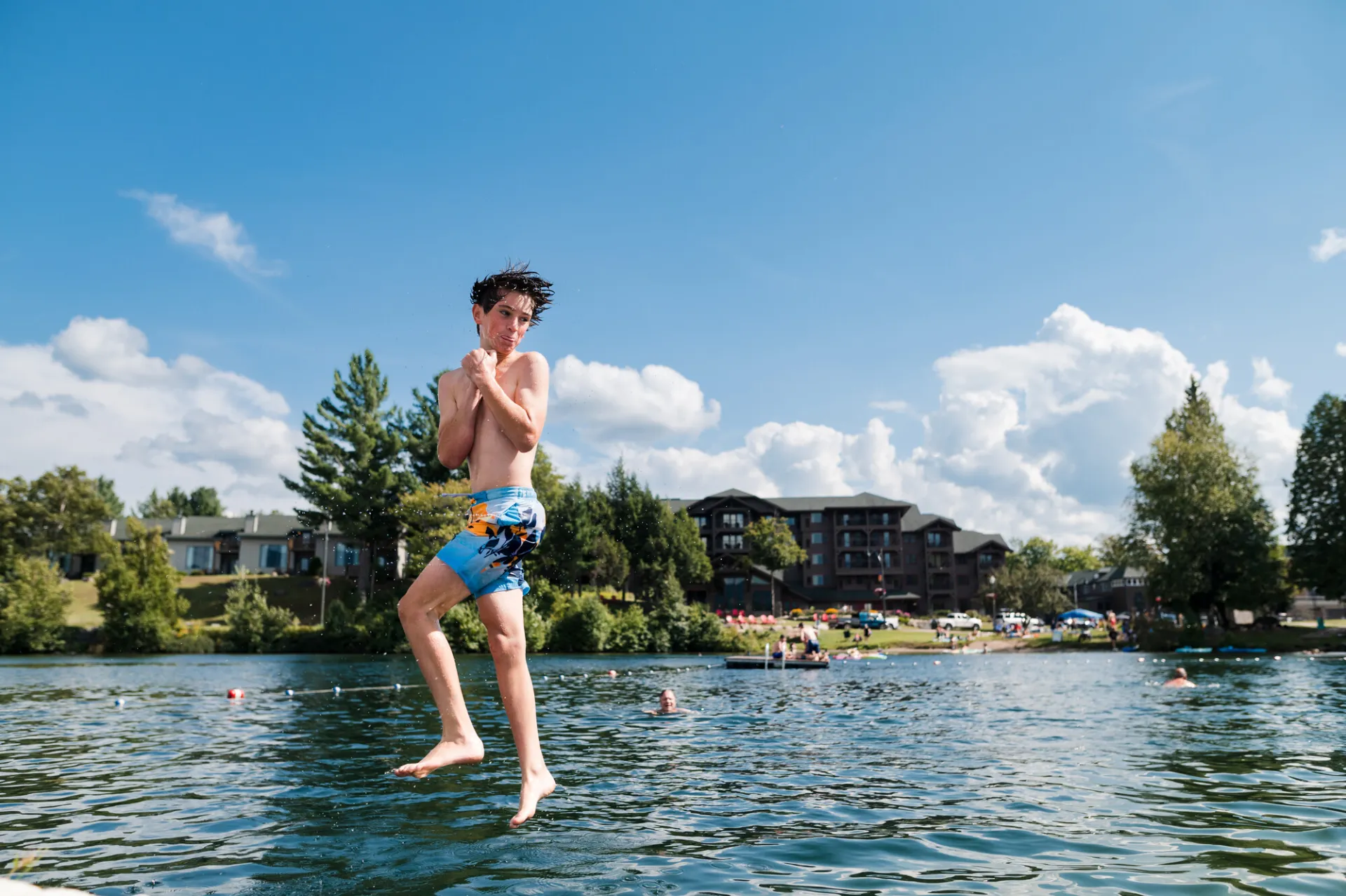 A boy in swim trunks spins in the air over a lake as he leaps into the water at a small beach.