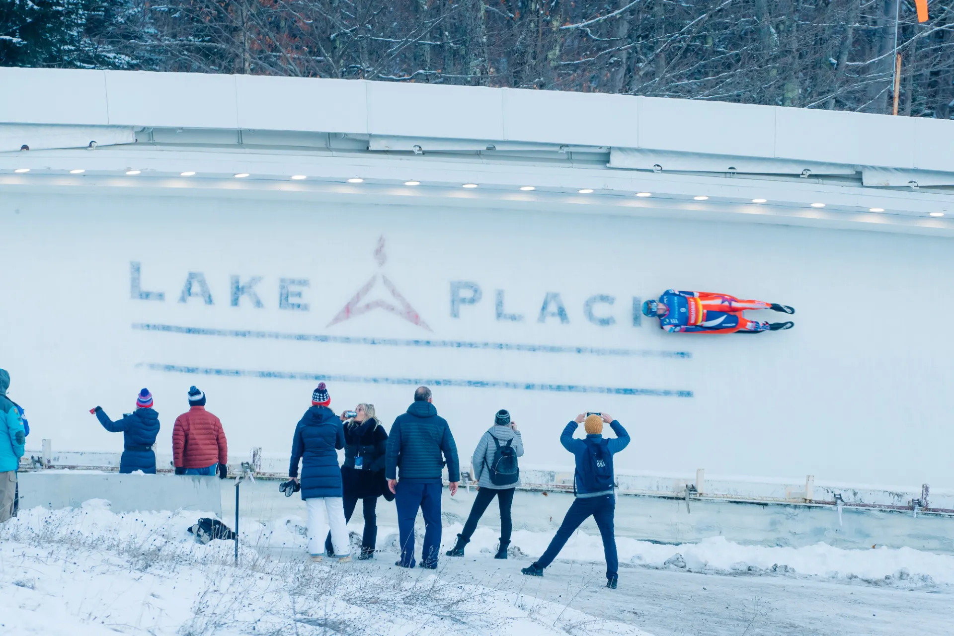 Luge racer rides down track with Lake Placid in ice while crowd looks on