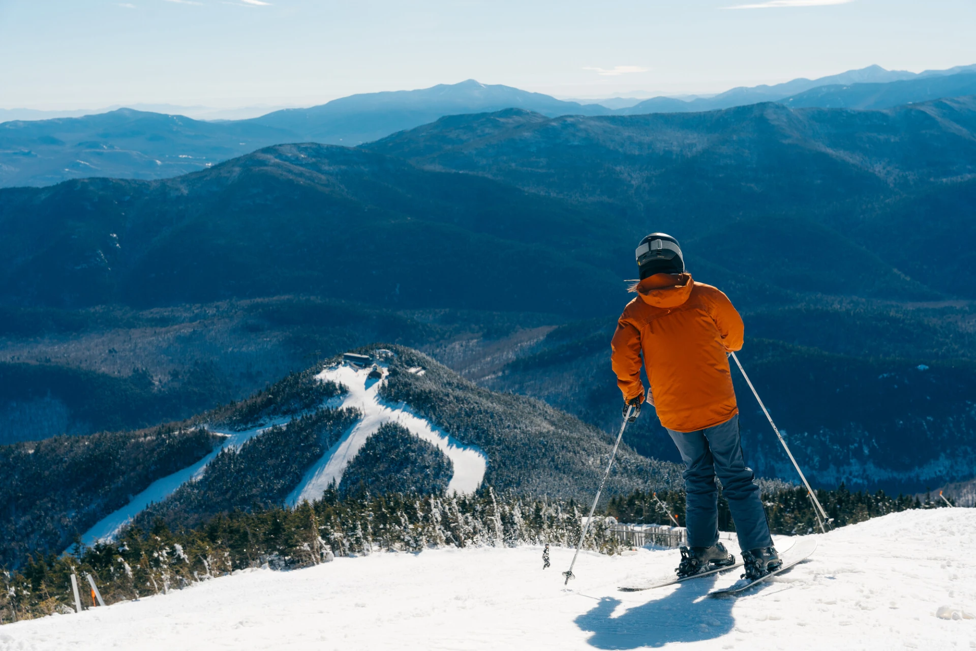 A skier in an orange jacket stands at the top of a snow-covered mountain, looking out over a range of blue-tinted peaks.