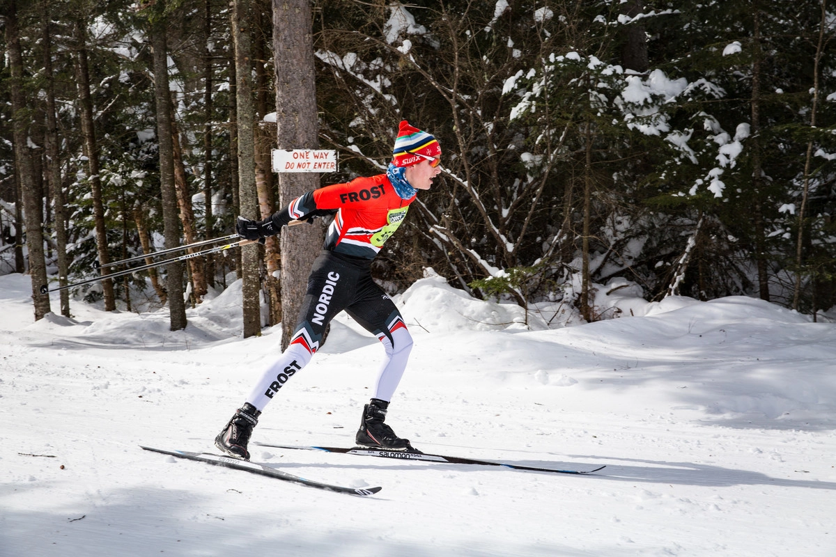 A single nordic skier passes by the camera from left to right, skiing on a snow-covered trail through a pine forest. They wear red, black, and white ski race gear and a yellow race bib on their chest