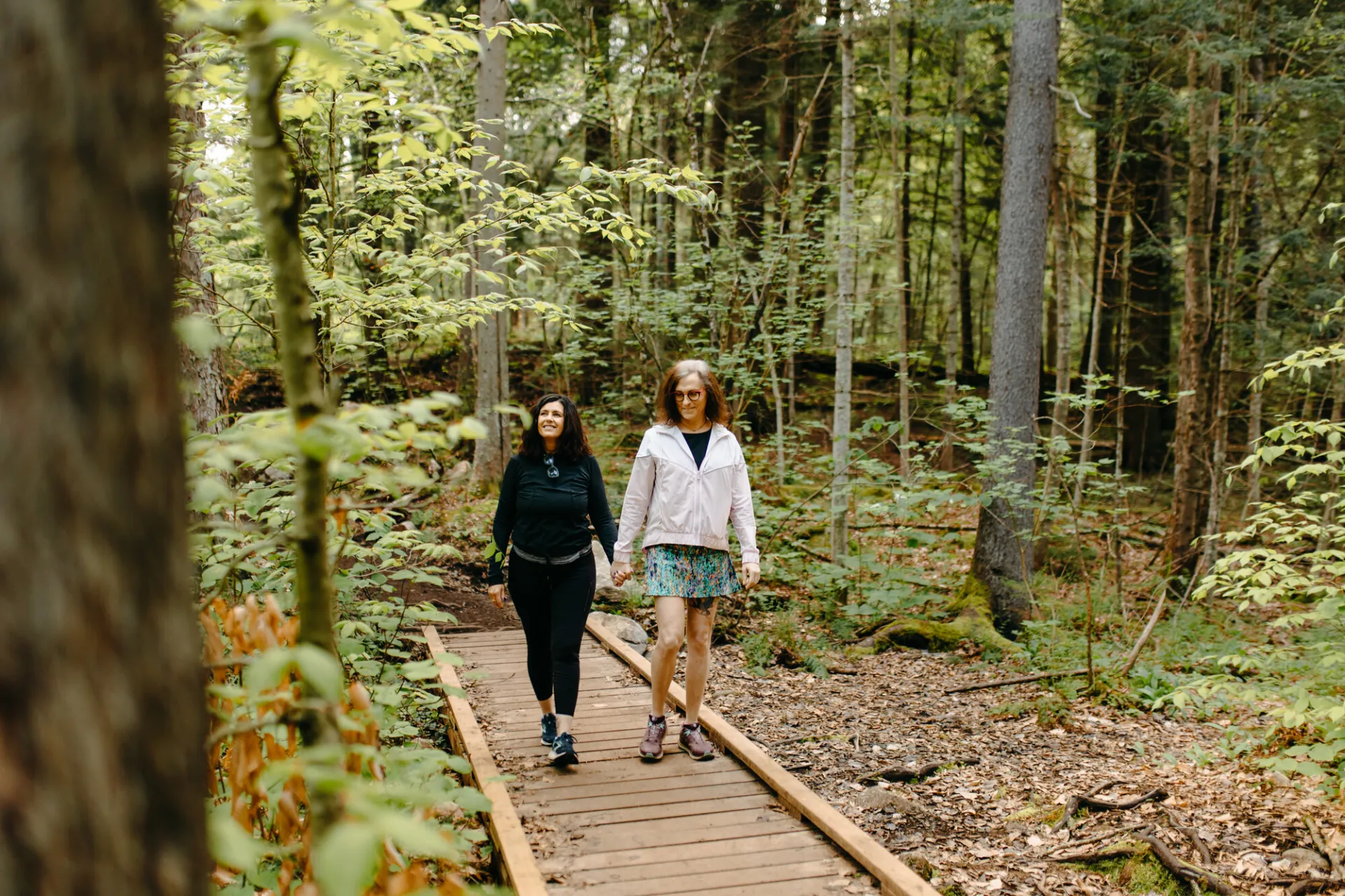 Two women walk on boardwalk path on trail through green woods