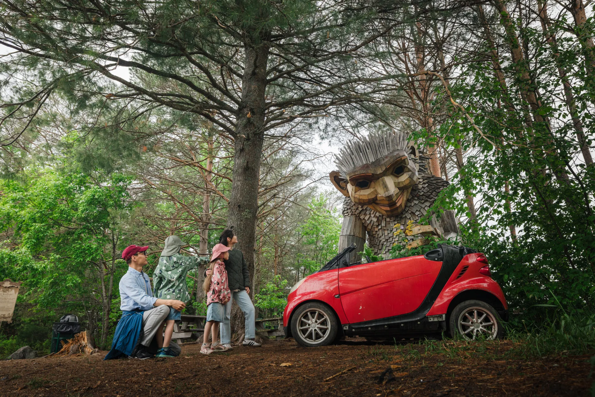 A family of two adults and two children examine a small red car underneath a large wooden troll.