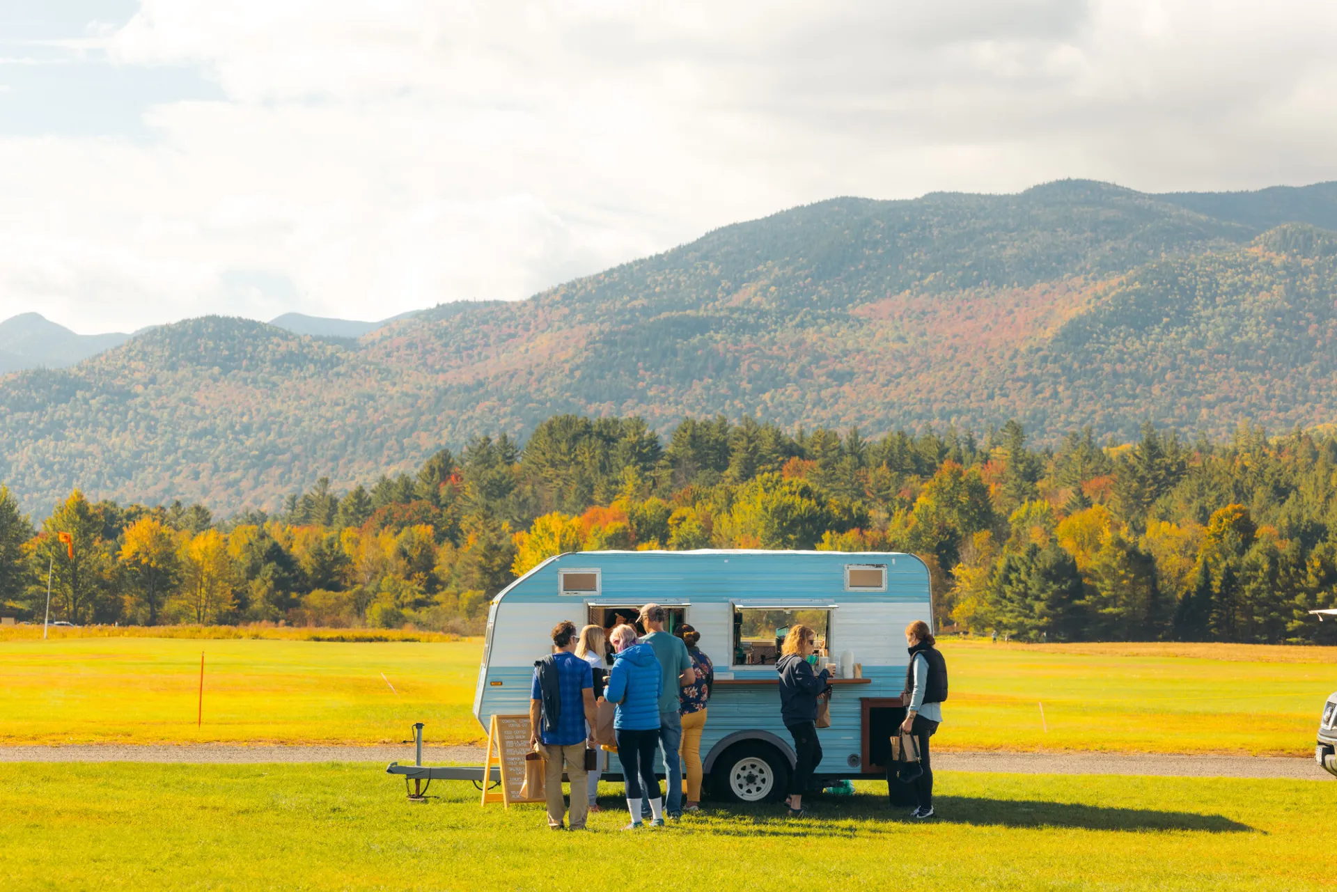 Blue and white camper trailer parked in field with mountains in background and people ordering coffee at window
