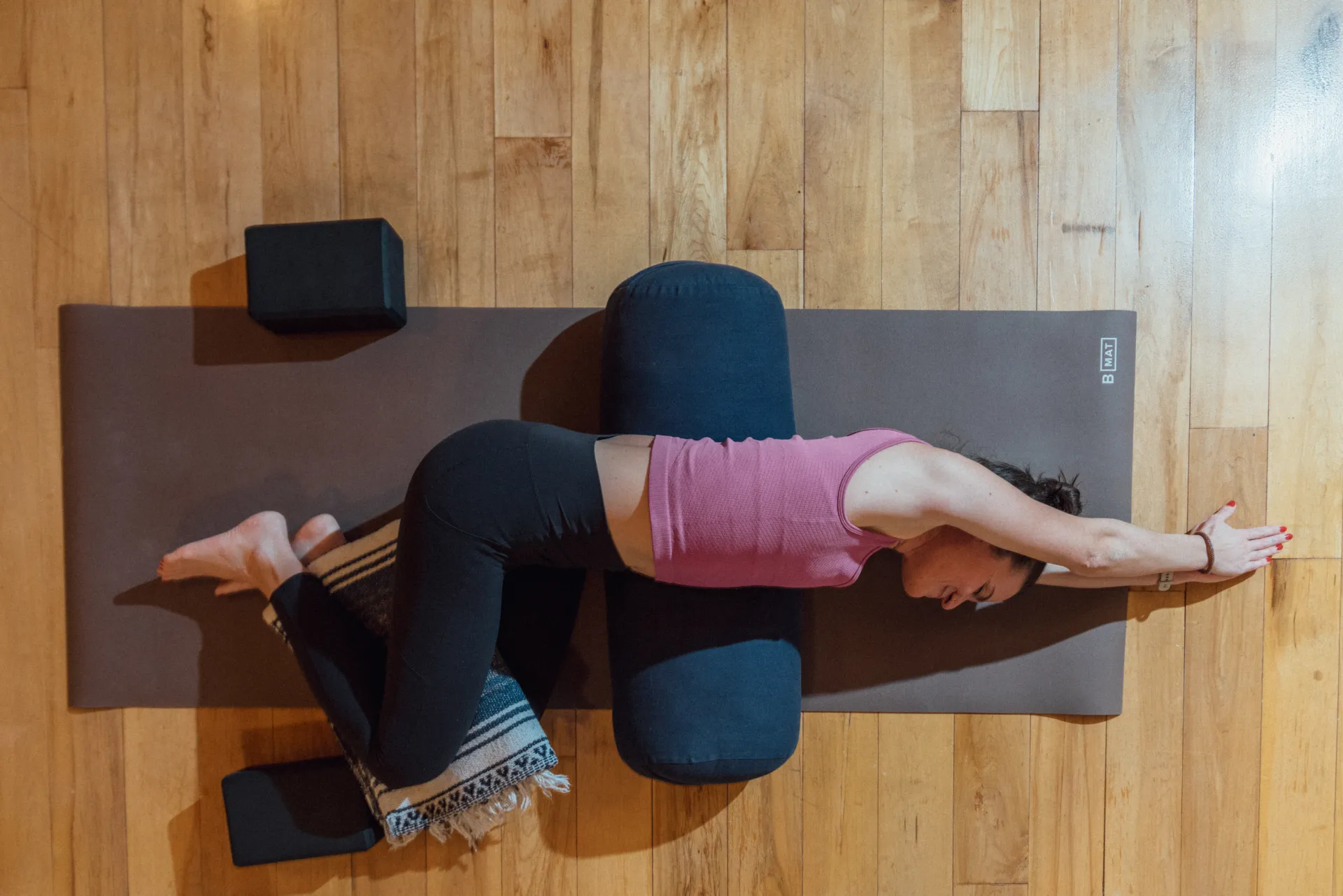 Woman in pink tank top and black yoga pants lays on side on black yoga mat on wooden floor with legs bent, a blanket between her knees, and her arms extended above her head