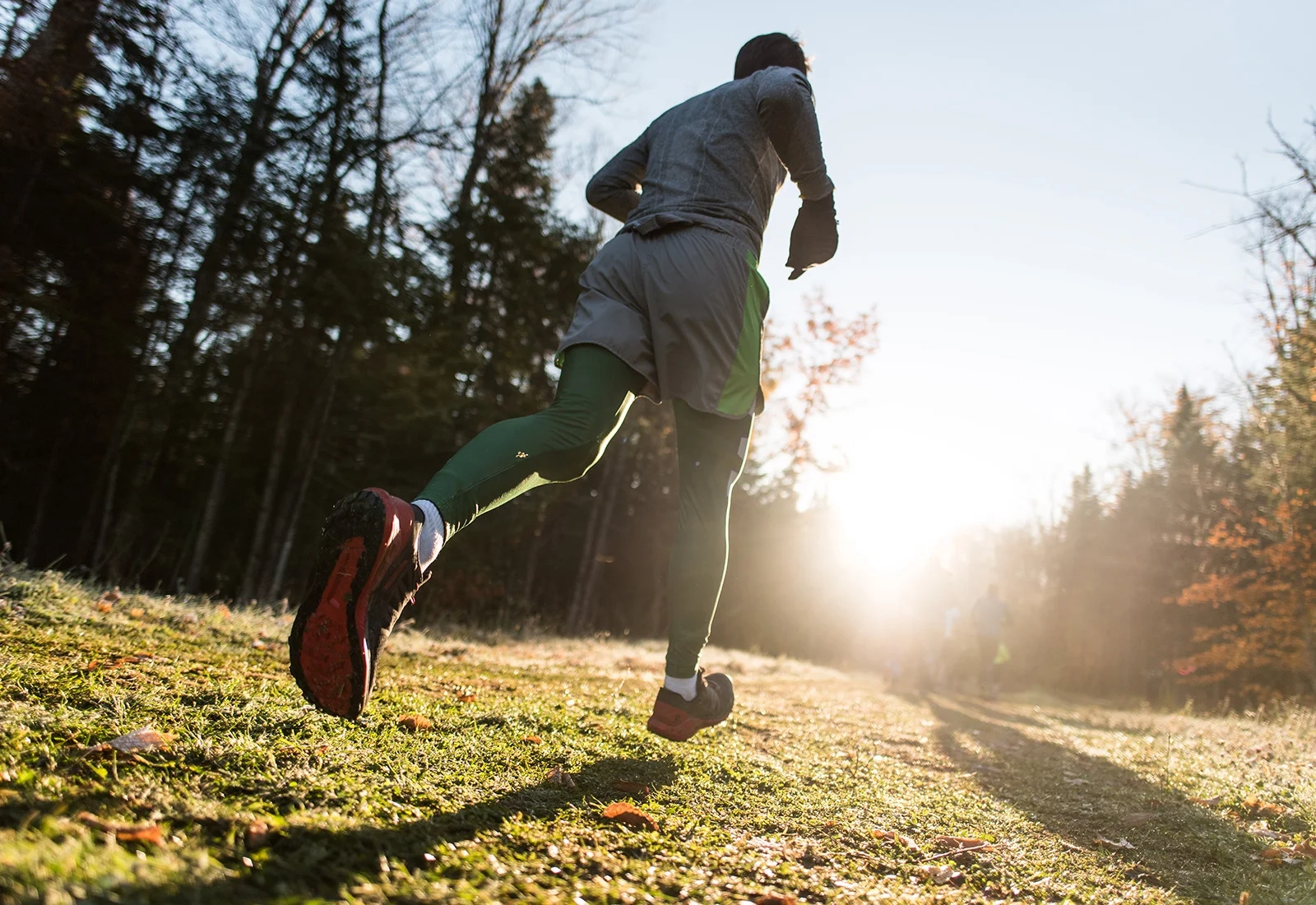 Close up of person's feet as they run on grassy path toward sunlight with bare and evergreen trees on sides of path