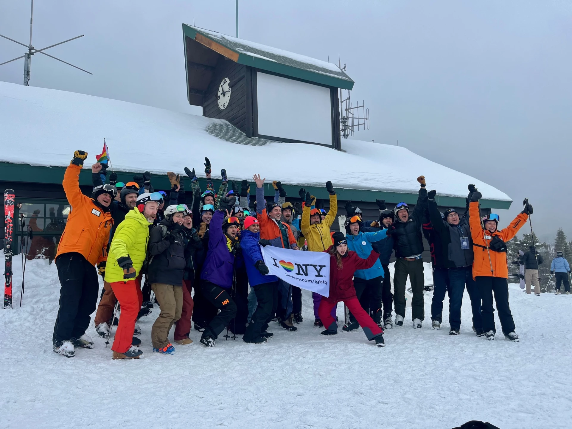 A large group of skiers and snowboarders in winter gear cheer and wave while holding an "I Love NY" LGBTQ+ pride flag.