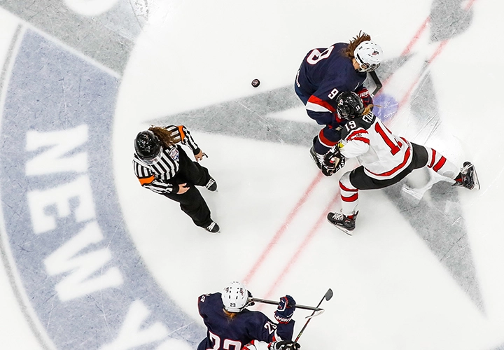Two female hockey players face off on ice with another playing and a referee looking on