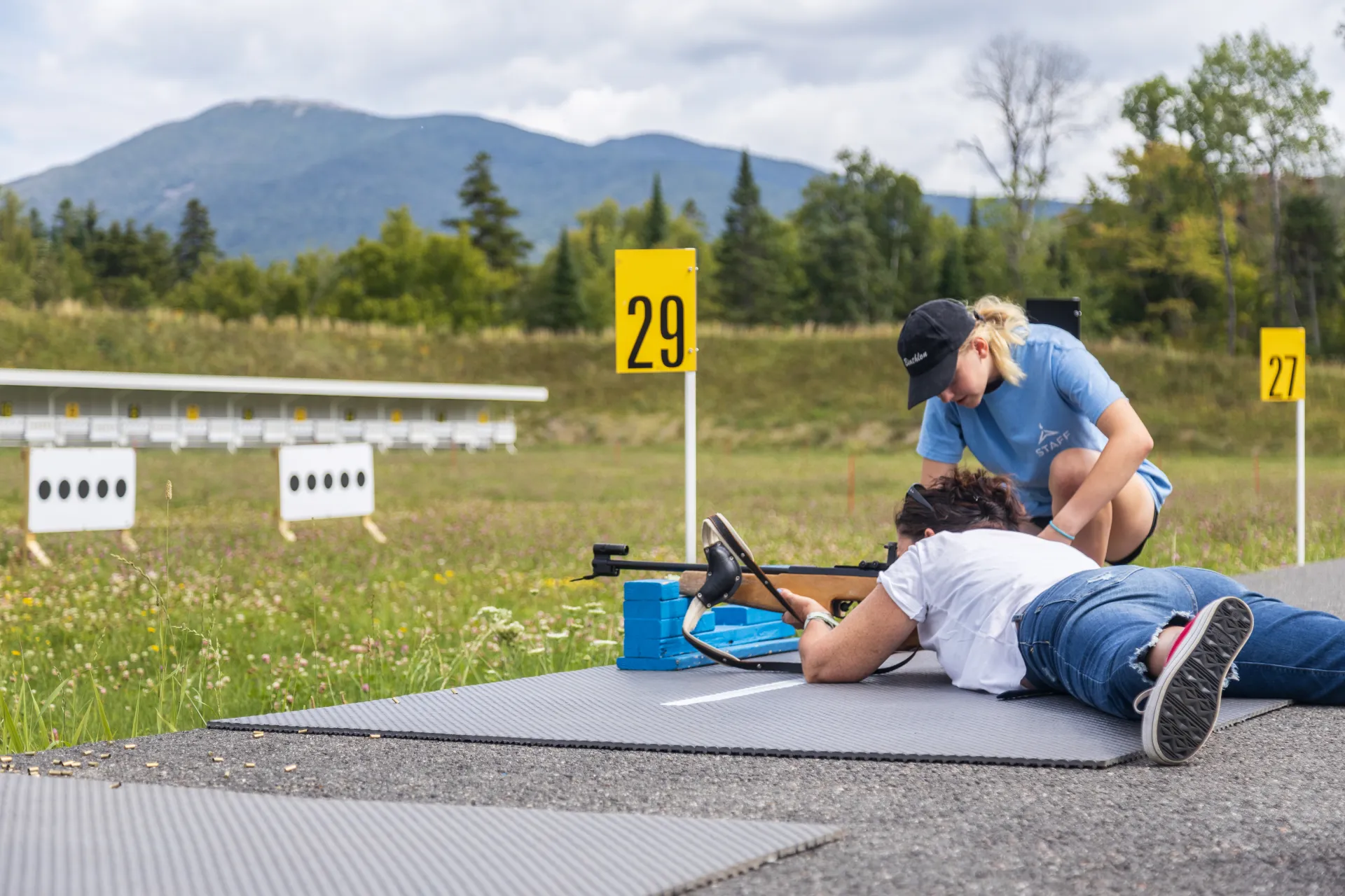 ORDA staff instructor kneels next to woman lying on biathlong shooting platform with biathlon rifle at range in summer