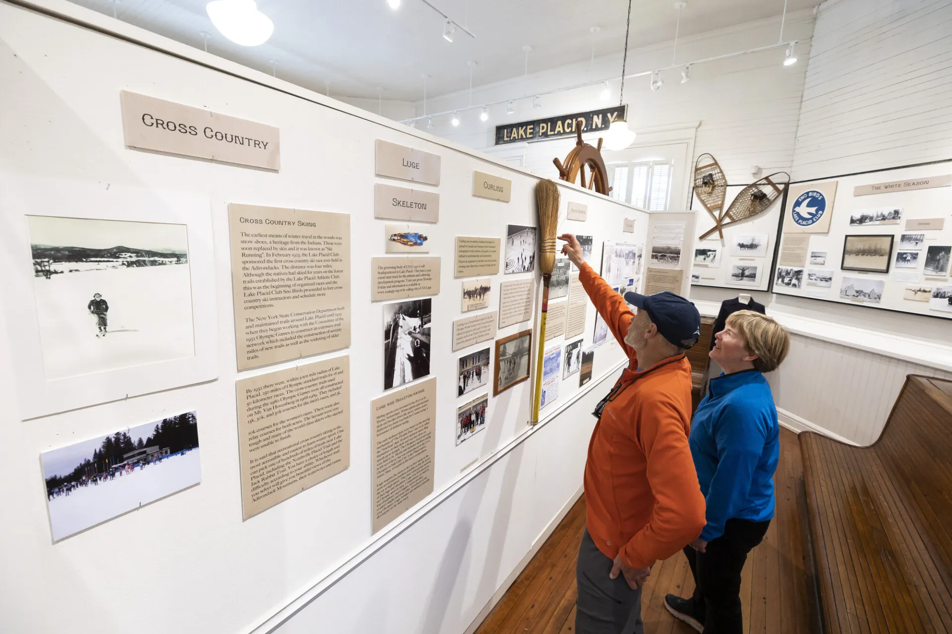 Senior man wearing orange sweater and hat stands next to senior blonde woman with blue jacket, the man pointing at a straw broom that is part of a historical display wall