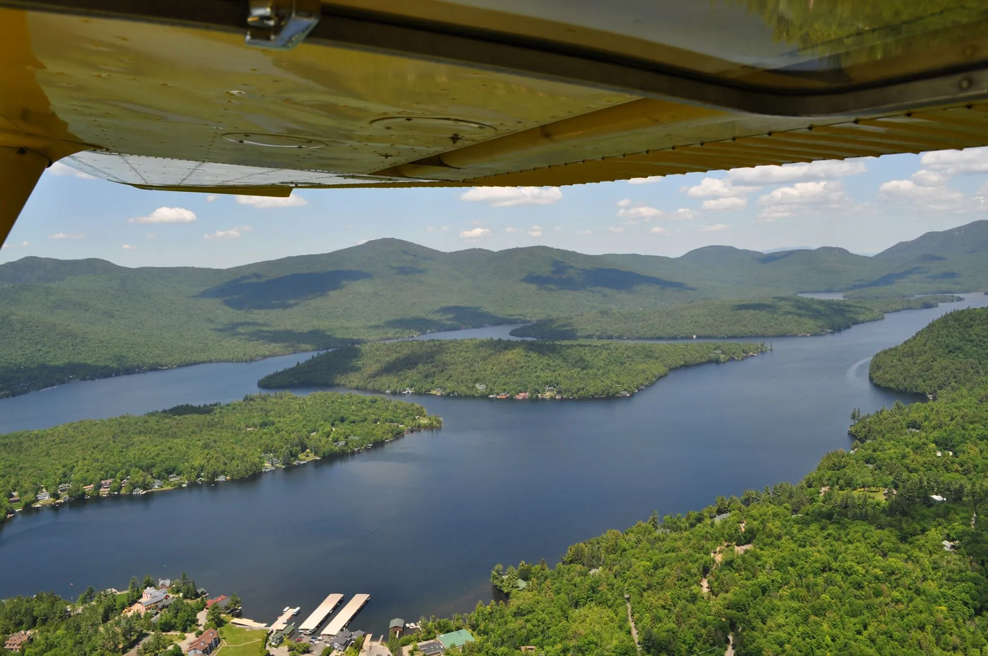 Looking down on green forests, a forested island, and blue lake from under a small yellow airplane wing.