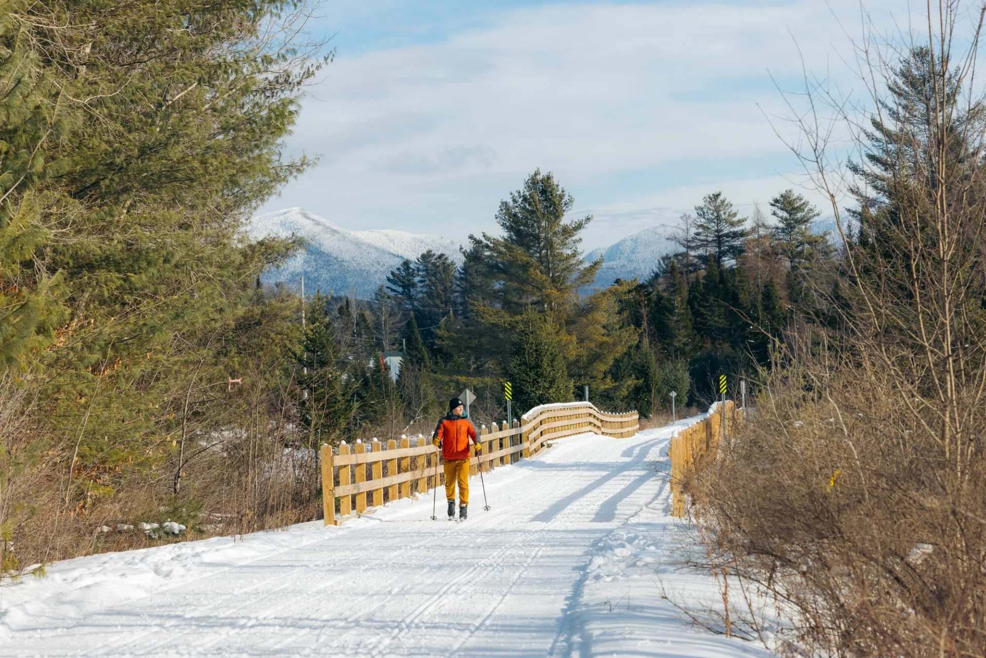 Person in red snow coat cross-country skis on Adirondack Rail Trail near bridge with snowy mountain seen in background