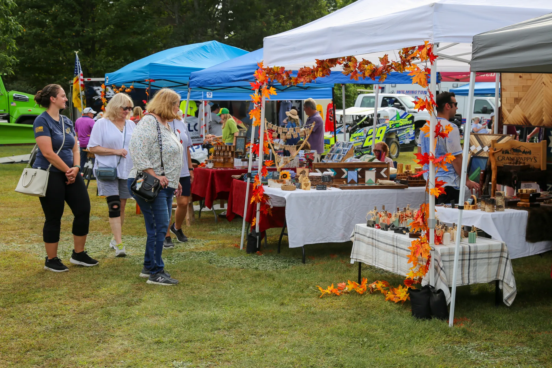 Three women look at the offerings at an outdoor vendor tent decorated with fall leaf garland