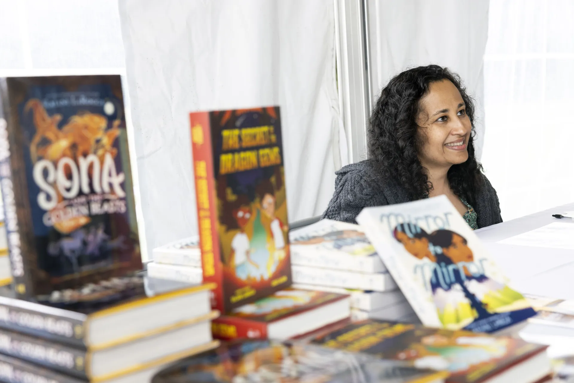Female author seen sitting and smiling at someone off camera from behind a stack and display of children's books
