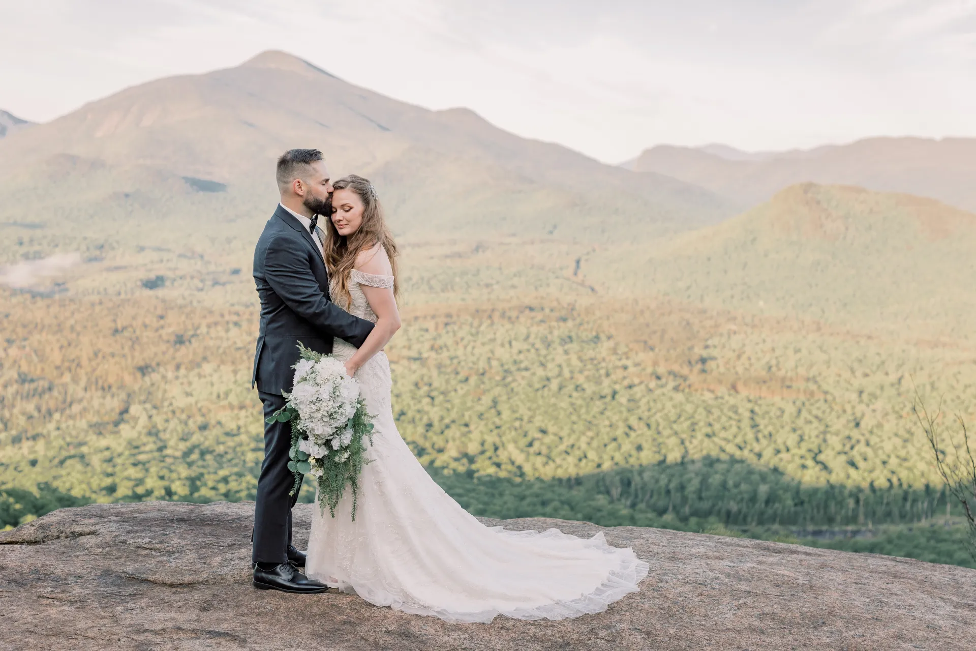 Groom kissses bride's forehead while she displays her bouquet on a rocky face with mountain views