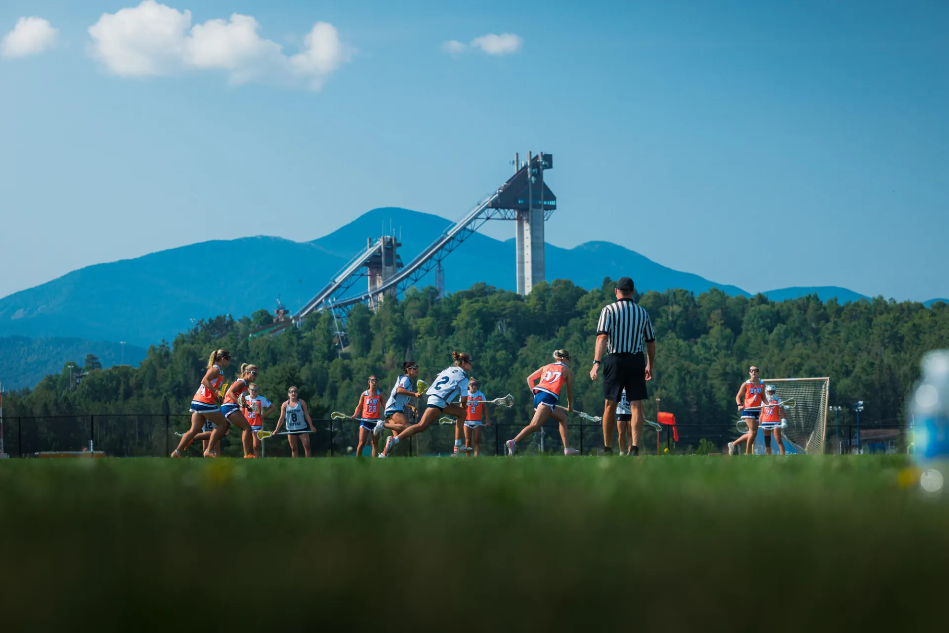 Two teams of women play lacrosse on field in front of mountains and ski jumps while a referee looks on