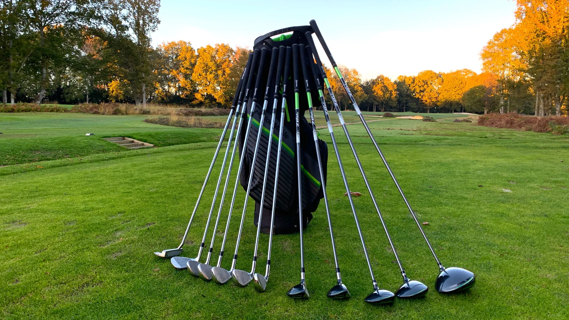 Set of golf clubs lined up leaning against golf bag standing independently on golf green with trees in background