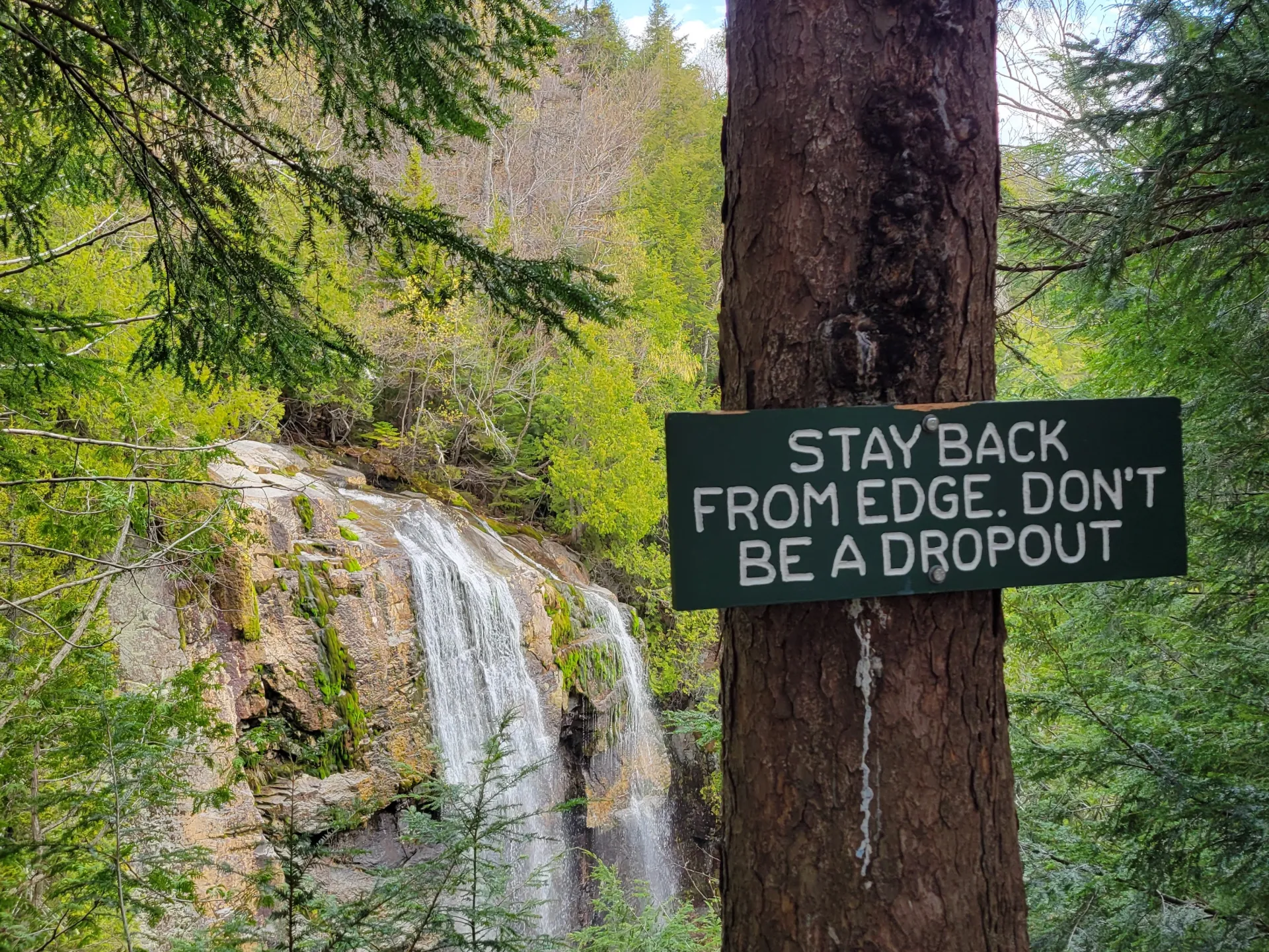A sign that says "Stay back from edge, don't be a drop out" in front of a waterfall