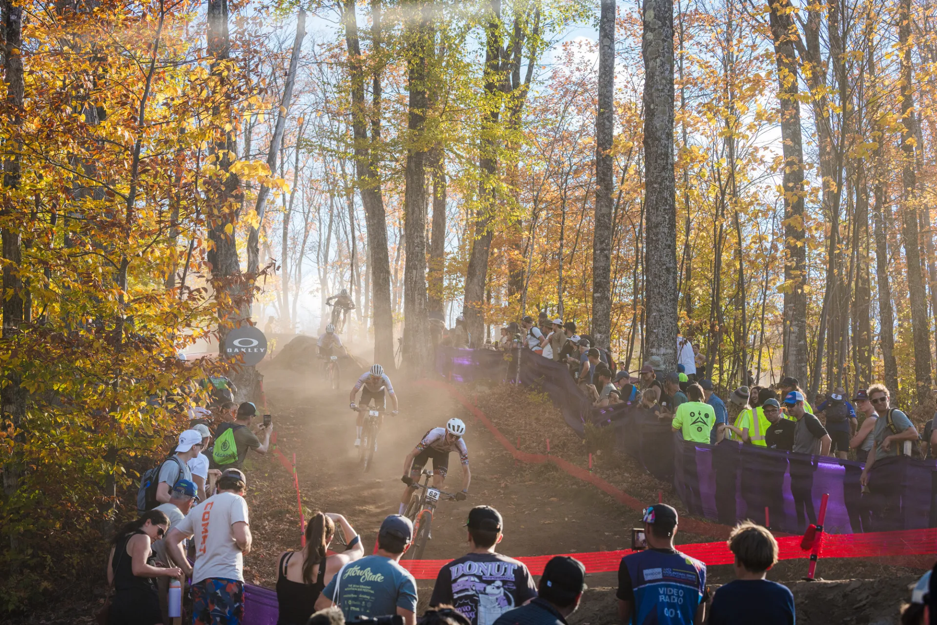 Mountain bike races ride down dirt course in forest with crowds flanking the course