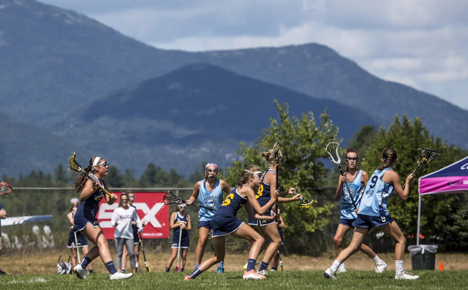 Two teams of young girls play lacrosse on green field with mountain backdrop.