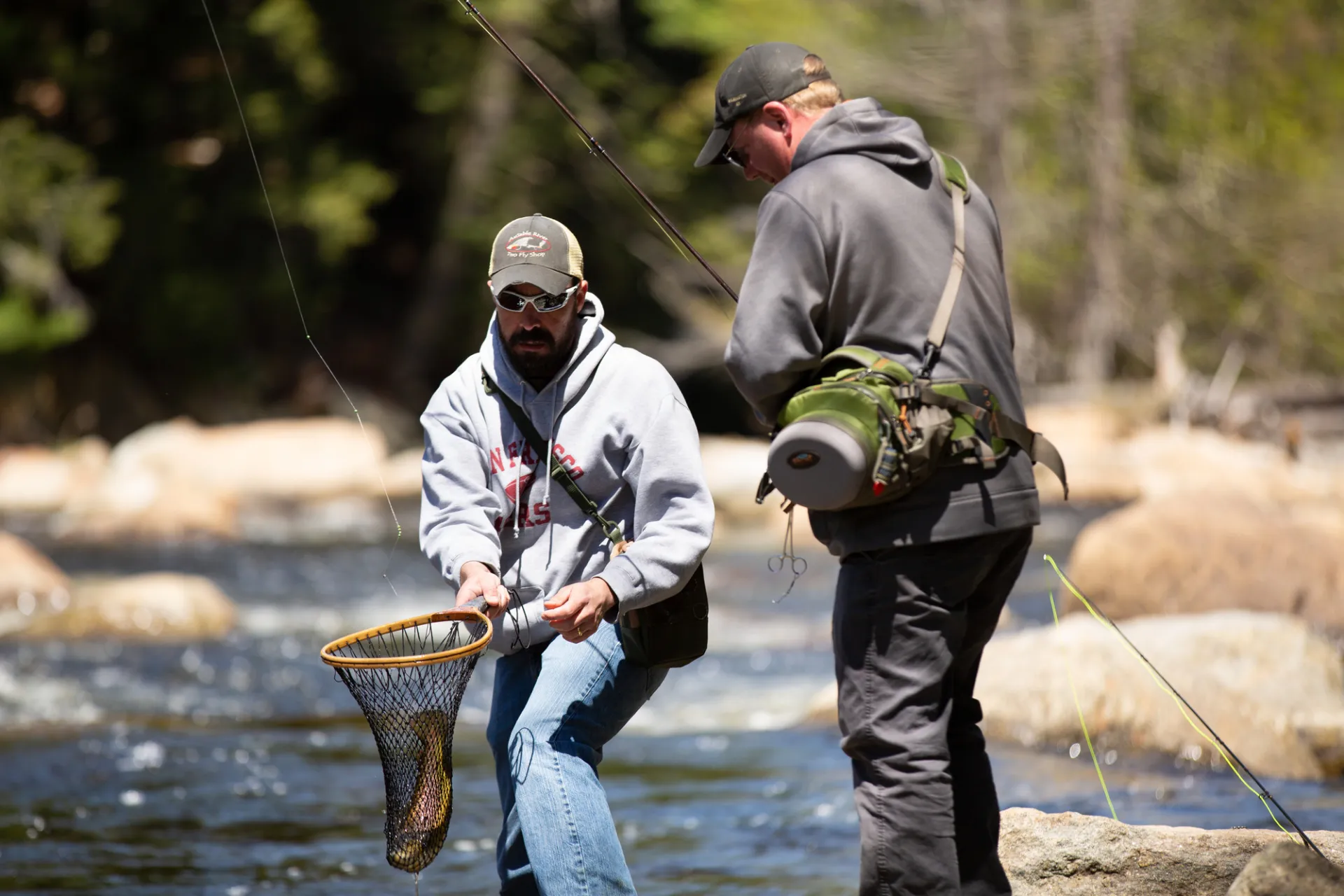 Two men fly fishing; one is reeling in a fish, while the other holds a net for the fish.