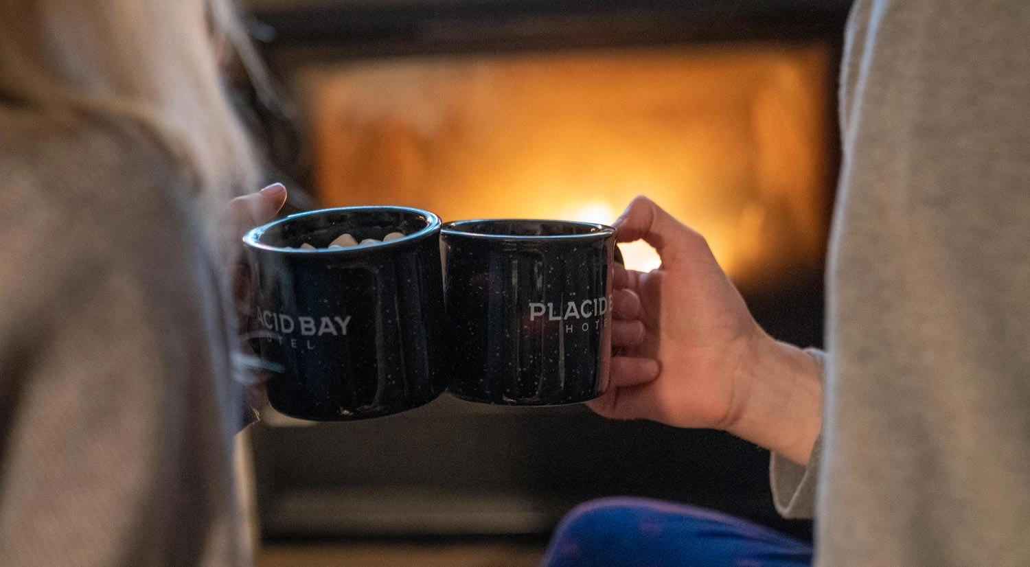 Two mugs of cocoa clinked together in front of a fire.