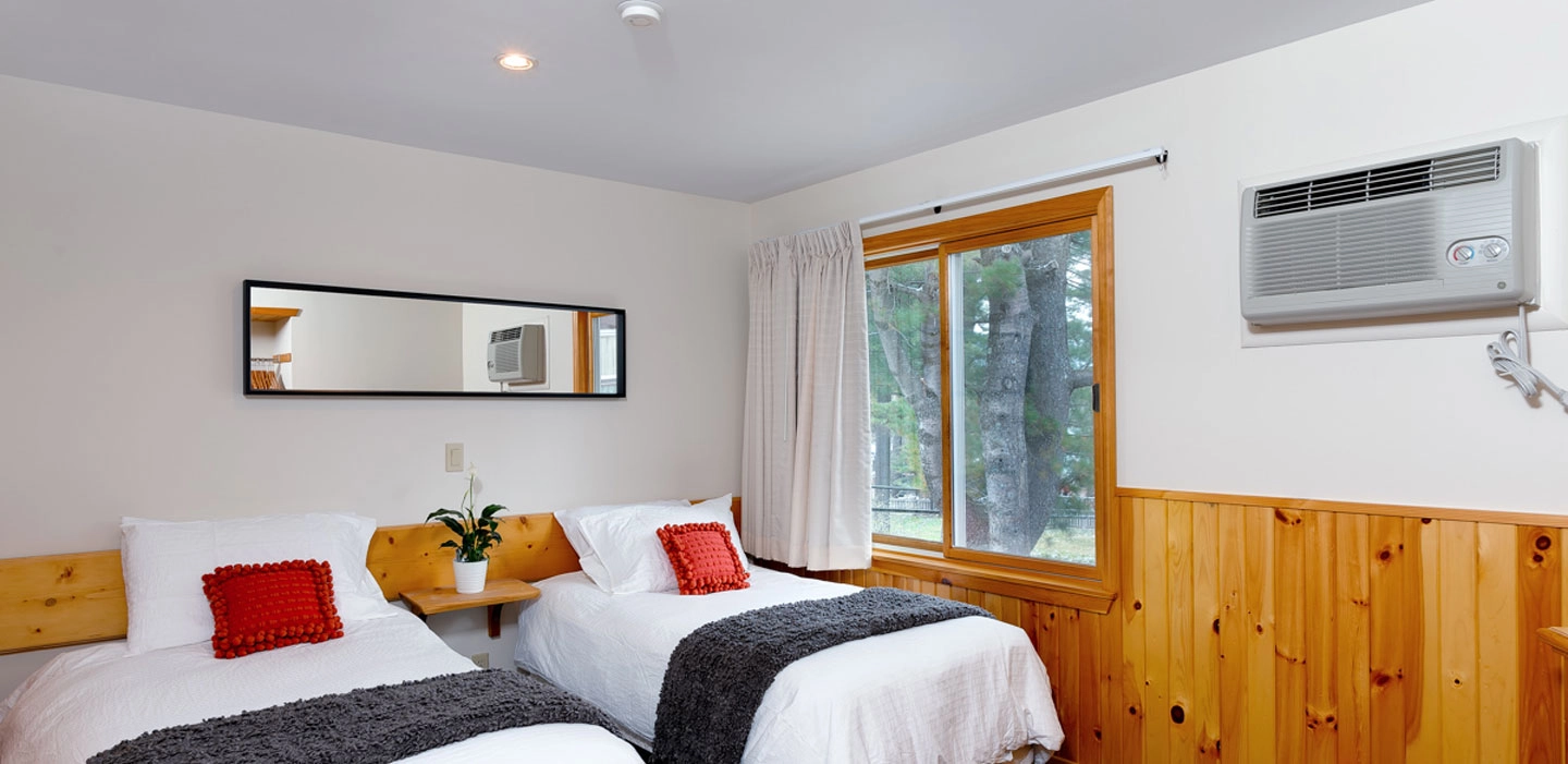 A two bed room option with white linens and blue accents and pine trees out the window.