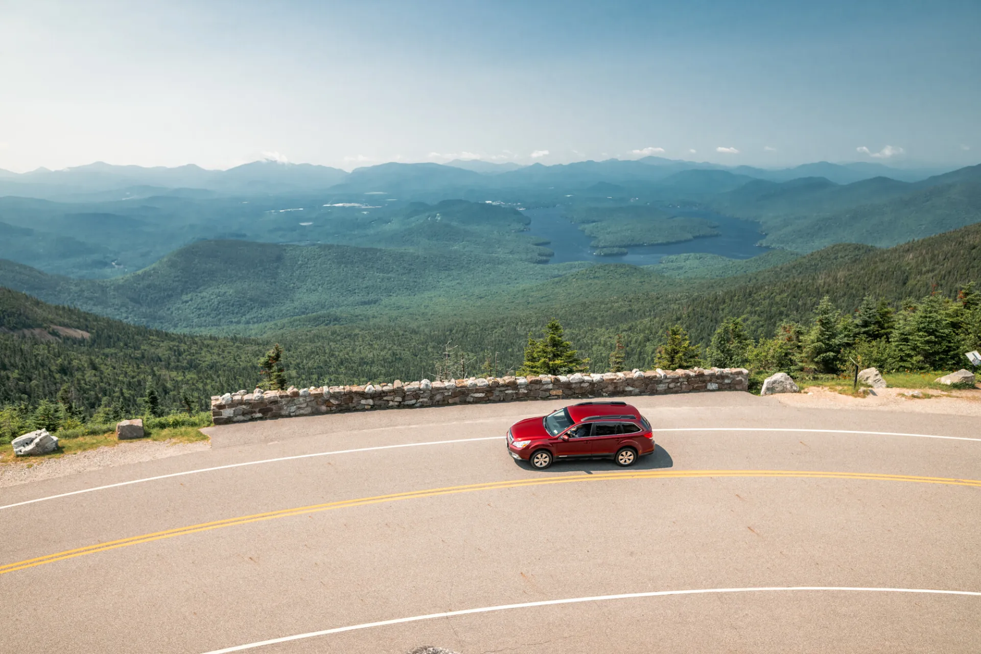 Red car drives on turn of high elevation highway road with mountain views and Lake Placid seen in the distance