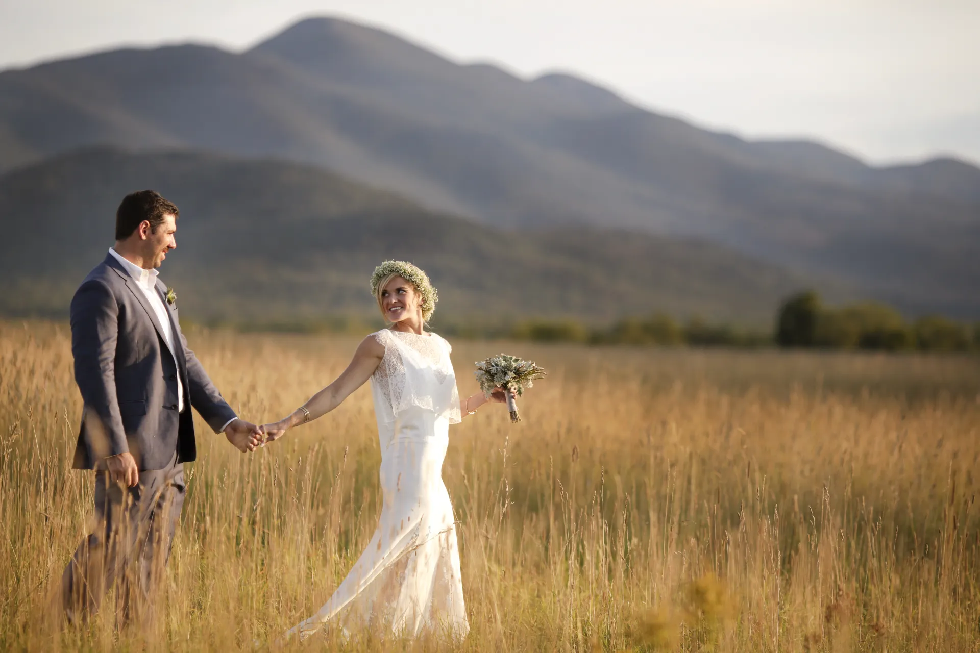 Bride and groom walk through a golden grass field with a mountain backdrop
