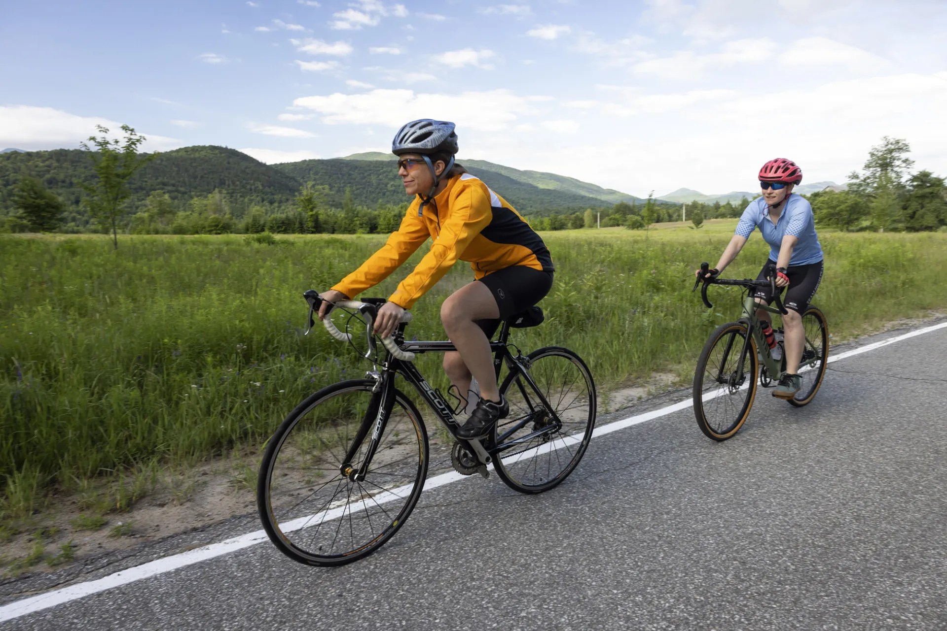 Cyclist wearing helmet and yellow and black rides single file ahead of second cyclist in light blue on paved road with green field and distant mountains in background