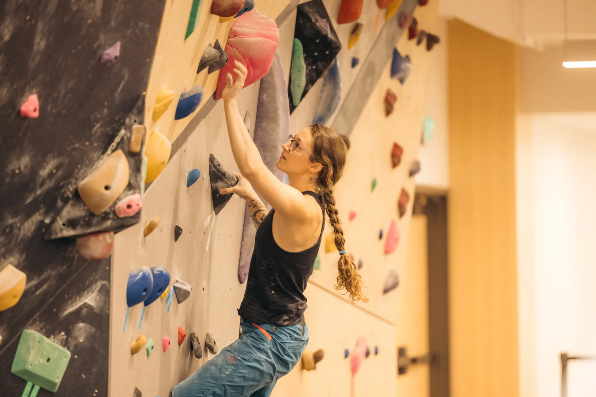 Female-presenting climber reaches for a hold on an indoor bouldering wall