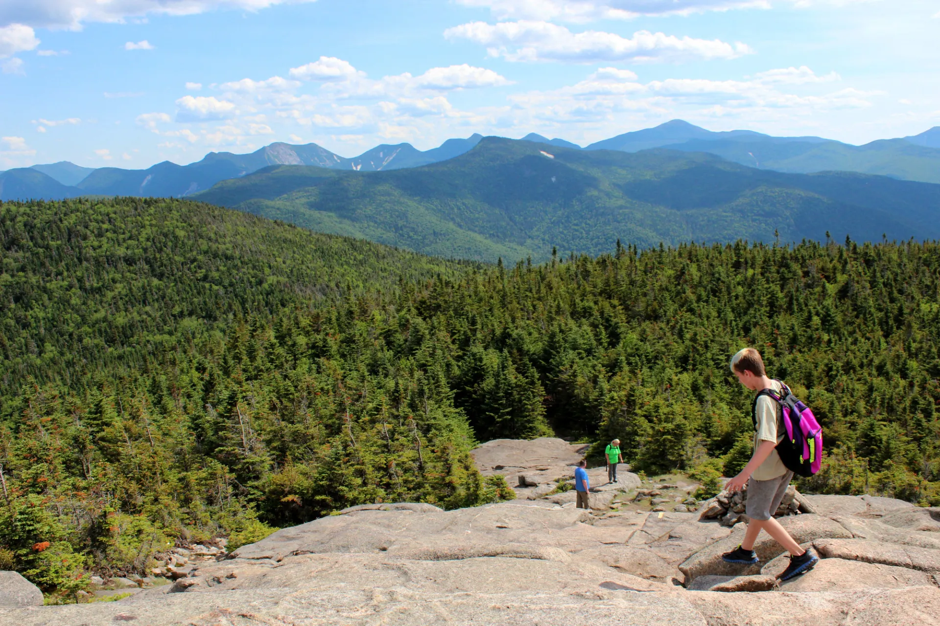 Views from the top of a hike with people hiking down a rock slab and mountain views in the background.
