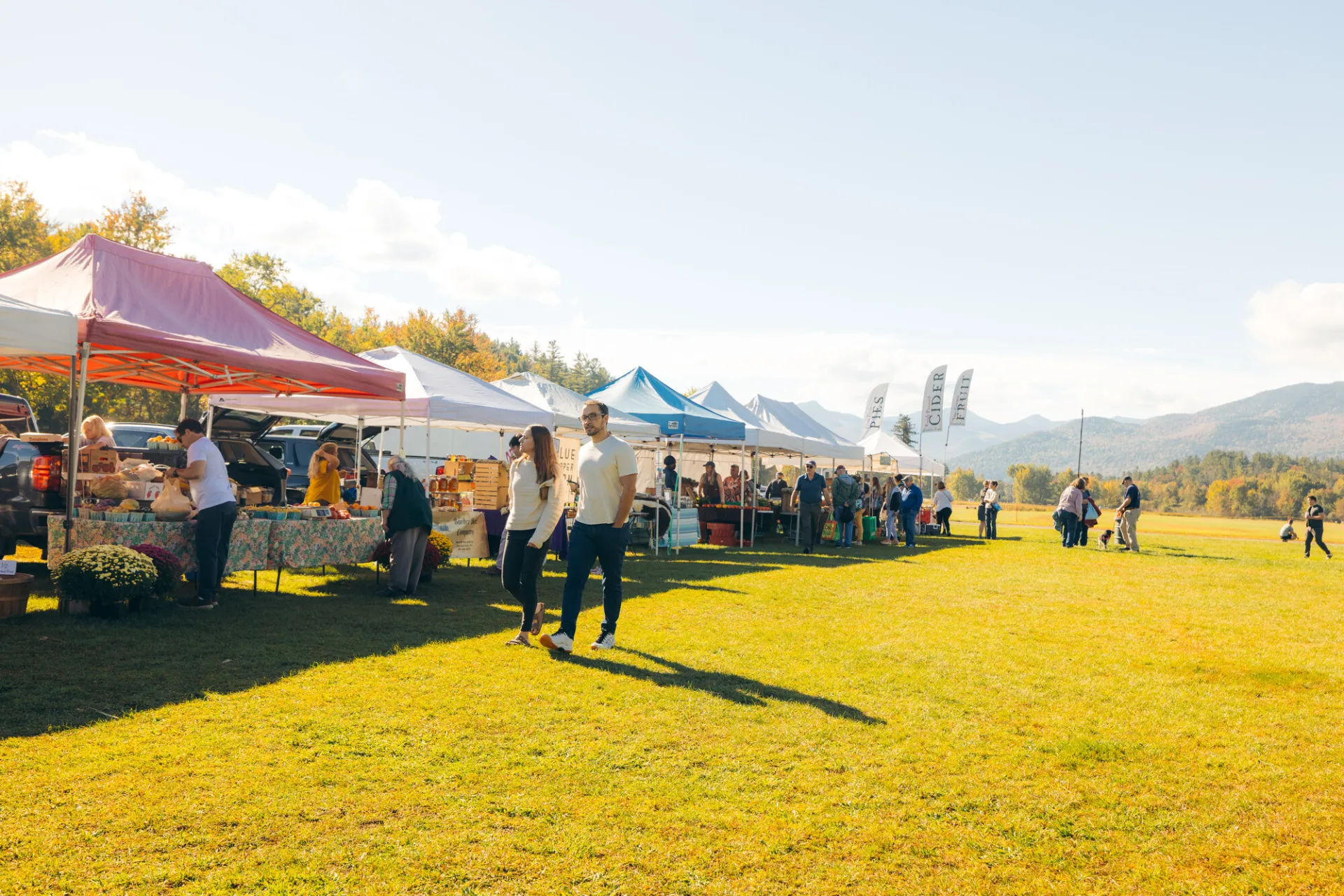 Couple walks past row of farmers market tents holding hands while other people browse vendors in background