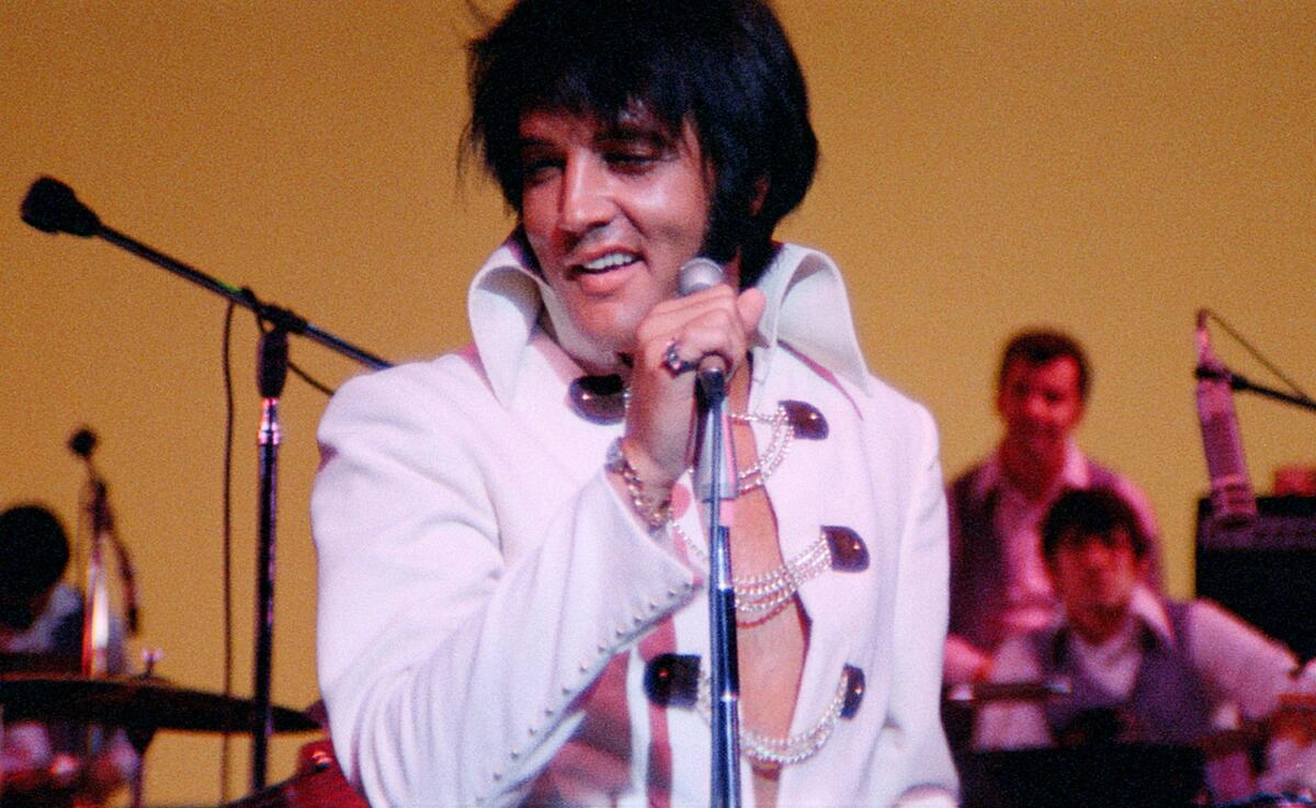 Still image of Elvis Presley in white costume on stage smiling behind microphone