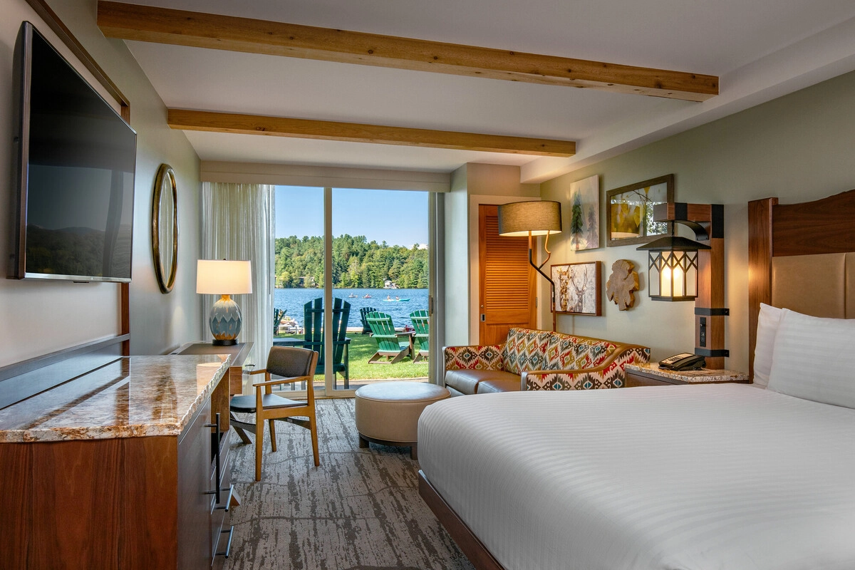 Hotel room with white bed and sitting area with sliding glass doors opening onto lakeside lawn with green Adirondack chairs