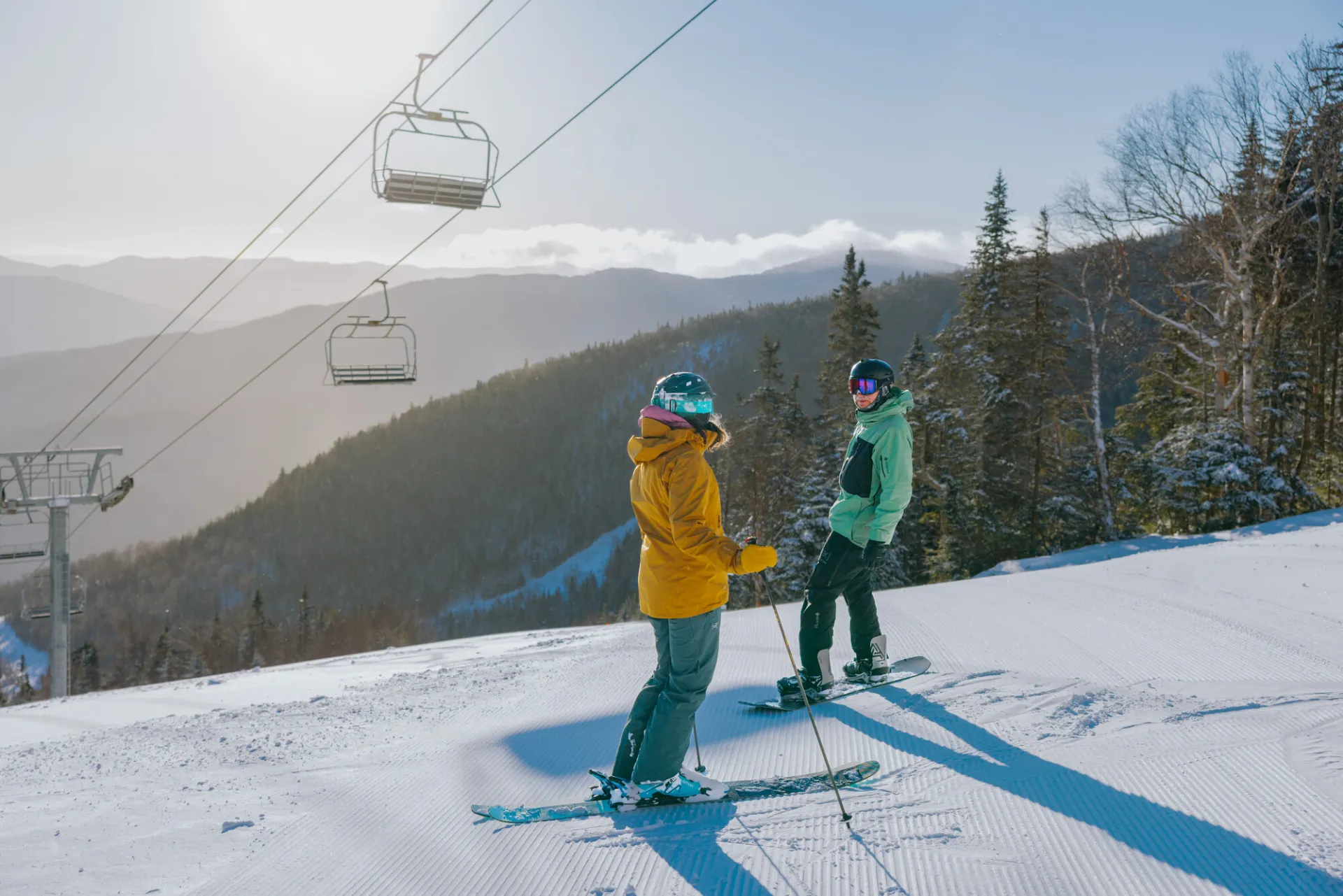A skier in a yellow jacket and a snowboarder in a green jacket pause on a groomed snowy slope under a chairlift.