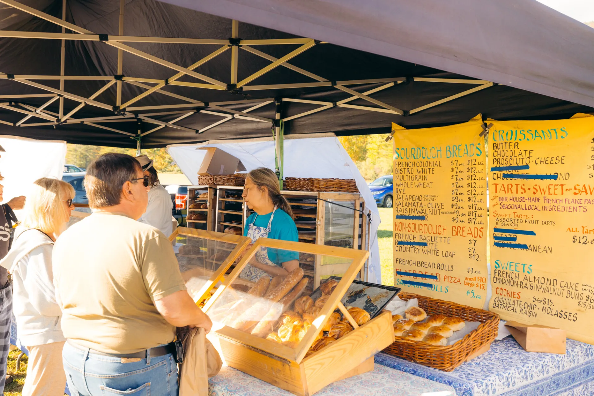 Person talks to seller over a table full of baked goods under outdoor farmers market tent