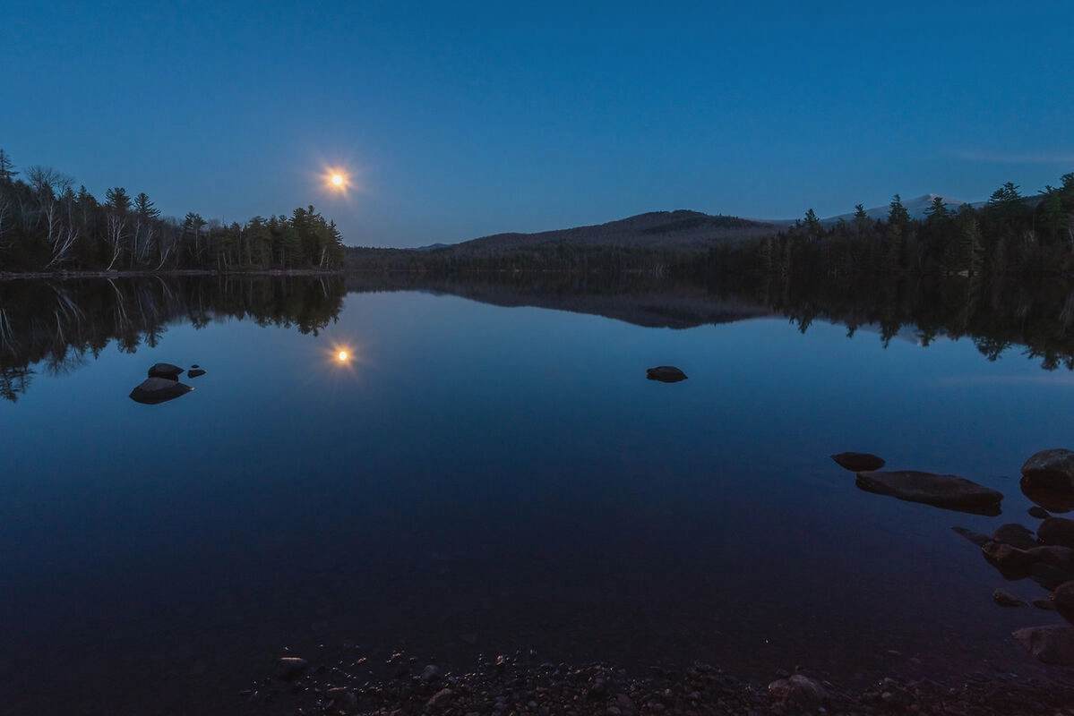 A tranquil lake at twilight reflects a full moon and the dark silhouettes of trees and mountains.  Several dark rocks are visible near the shore.