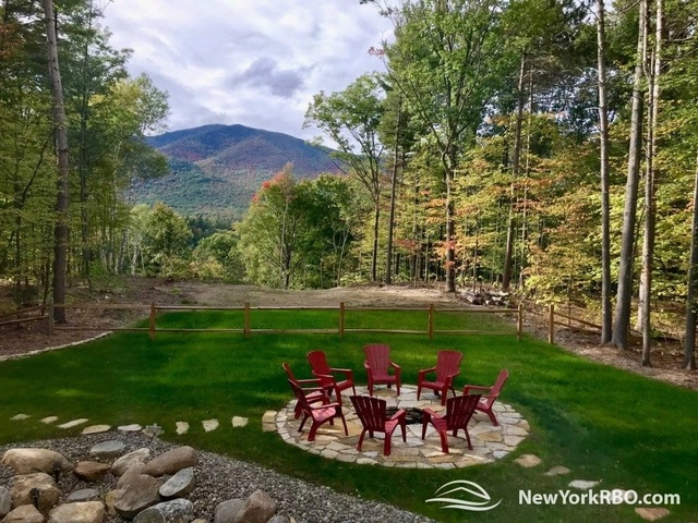 A lush and expansive green lawn with red adirondack style chairs arranged on a patio with fire pit and views of the mountains beyond the trees.