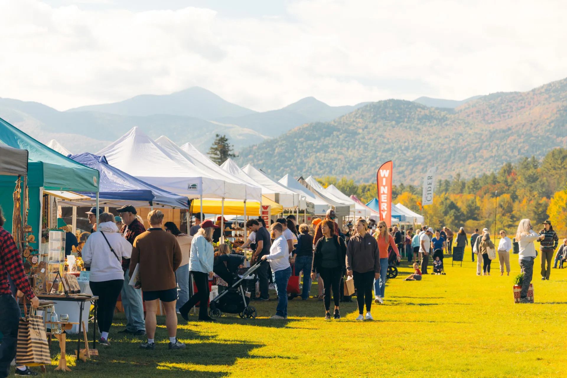 Crowd seen shopping and browsing rows of tents at outdoor farmers market in fall with mountains in background showing fall colors