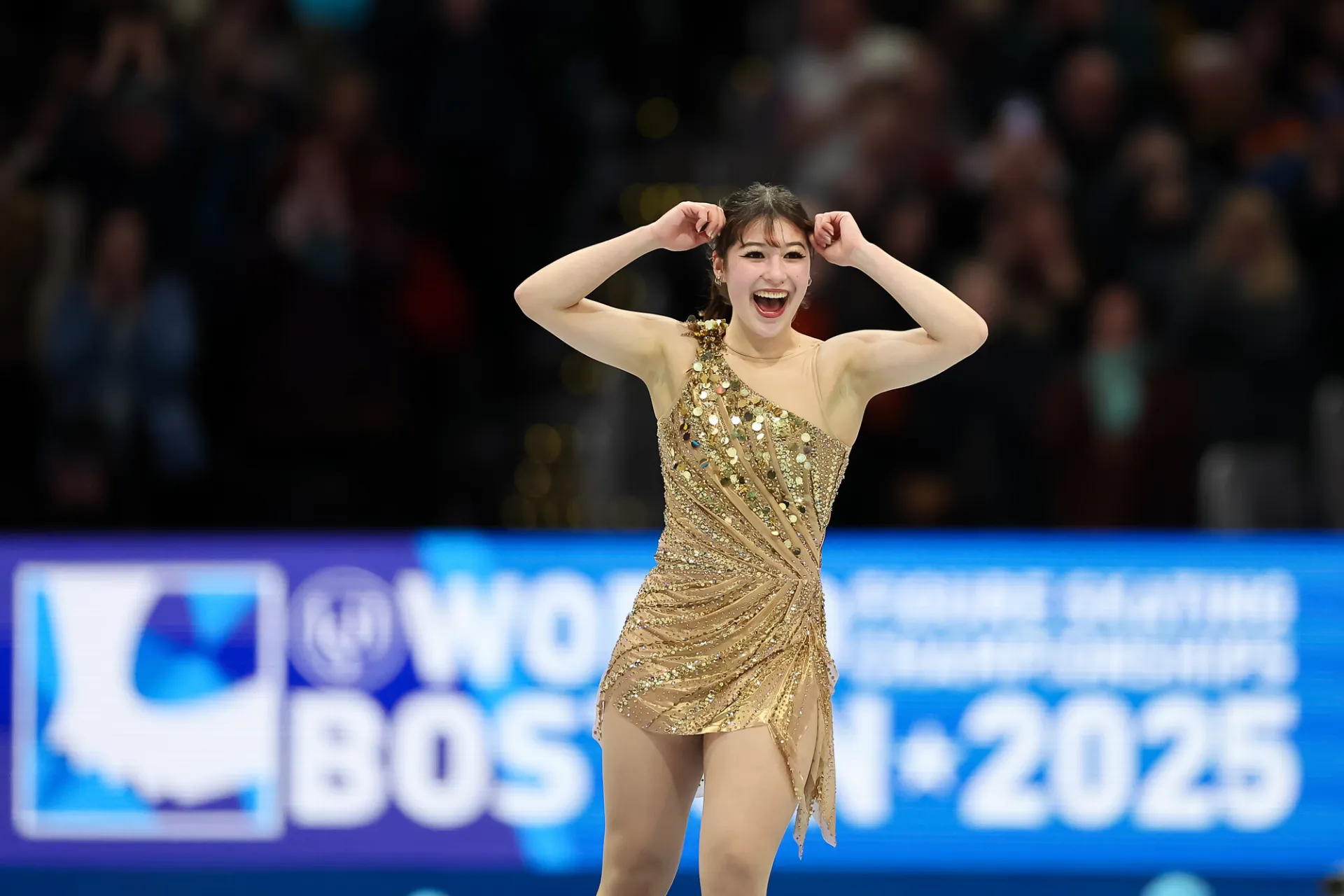 Woman in a gold dress smiles and raises arm in celebration while performing on ice at figure skating competition
