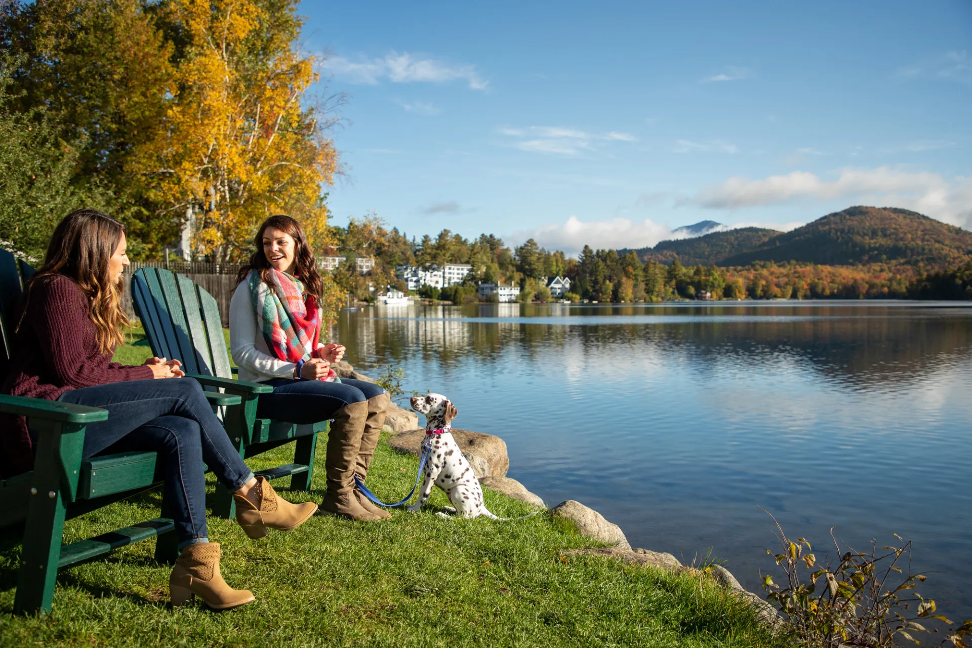 Two women and a dog sit in Adirondack chairs next to a calm lake with fall foliage around.