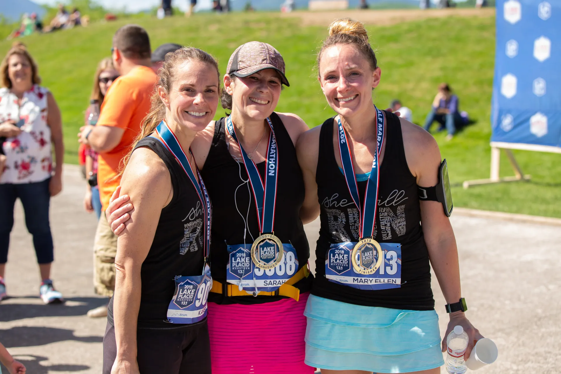 Three women wearing sports clothing and race numbers stand with arms around each other smiling wearing race finisher medals