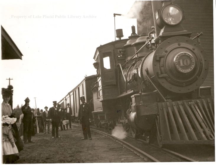 A vintage black and white image of a steam locomotive-powered train with passengers and officials on the ground nearby.