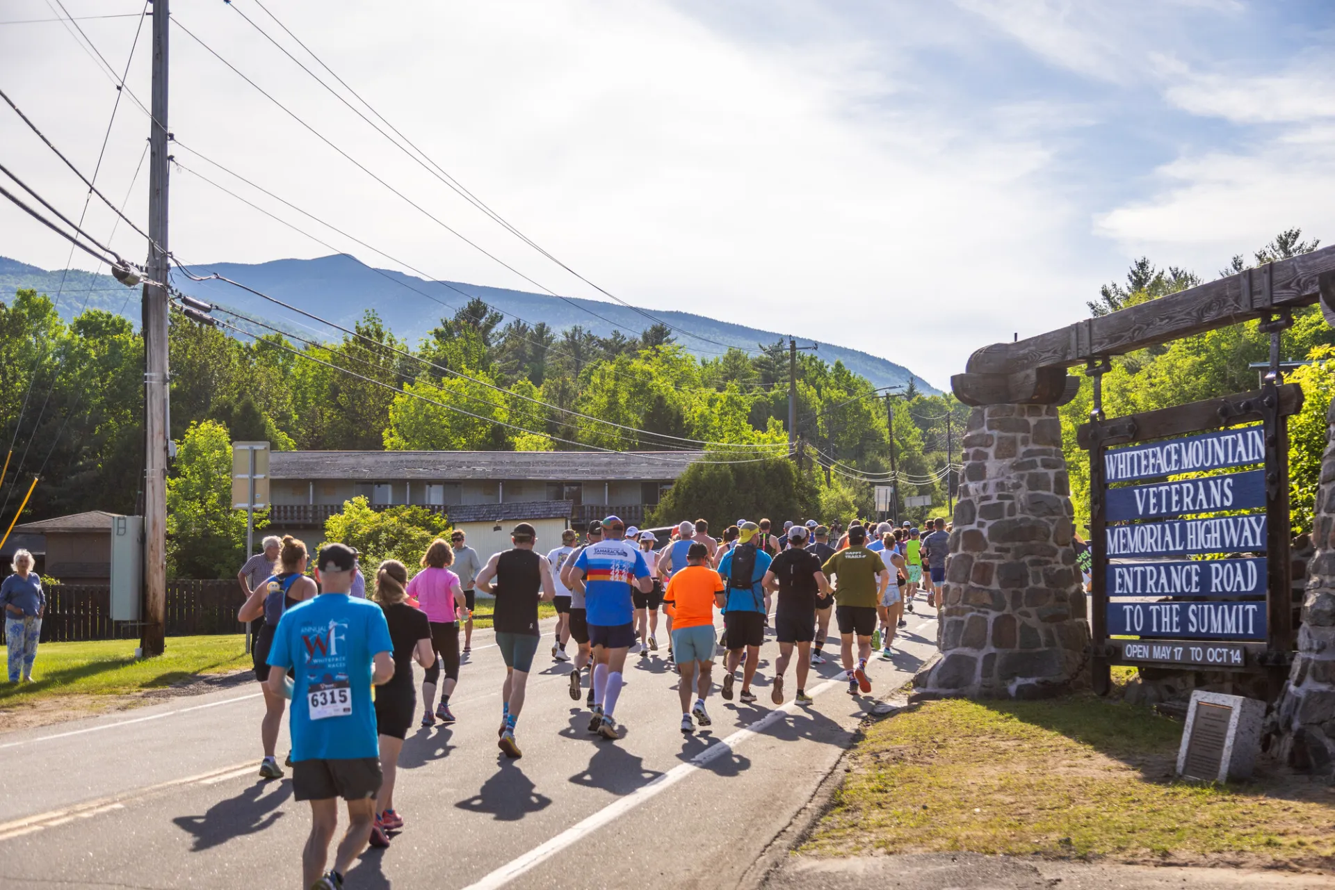 A large group of runners head down a road, passing a stone sign for the Whiteface Mountain Veterans Memorial Highway.