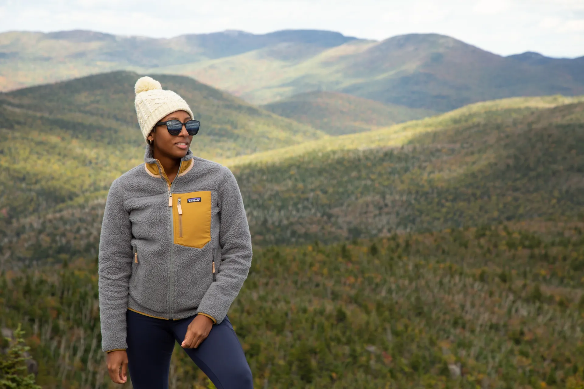 A hiker smiles facing the camera with a mountainous landscape in the background during late fall