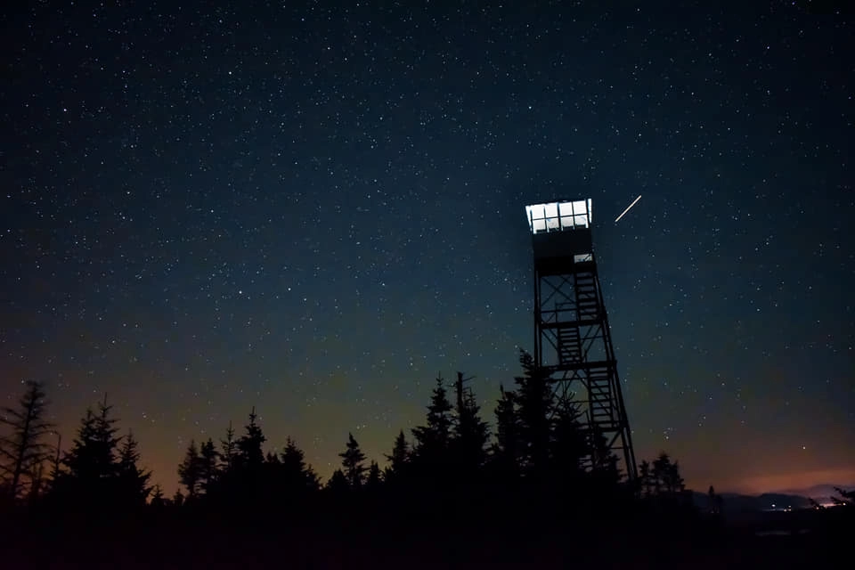 Silhouette of a fire tower at night under a starry sky with a shooting star.