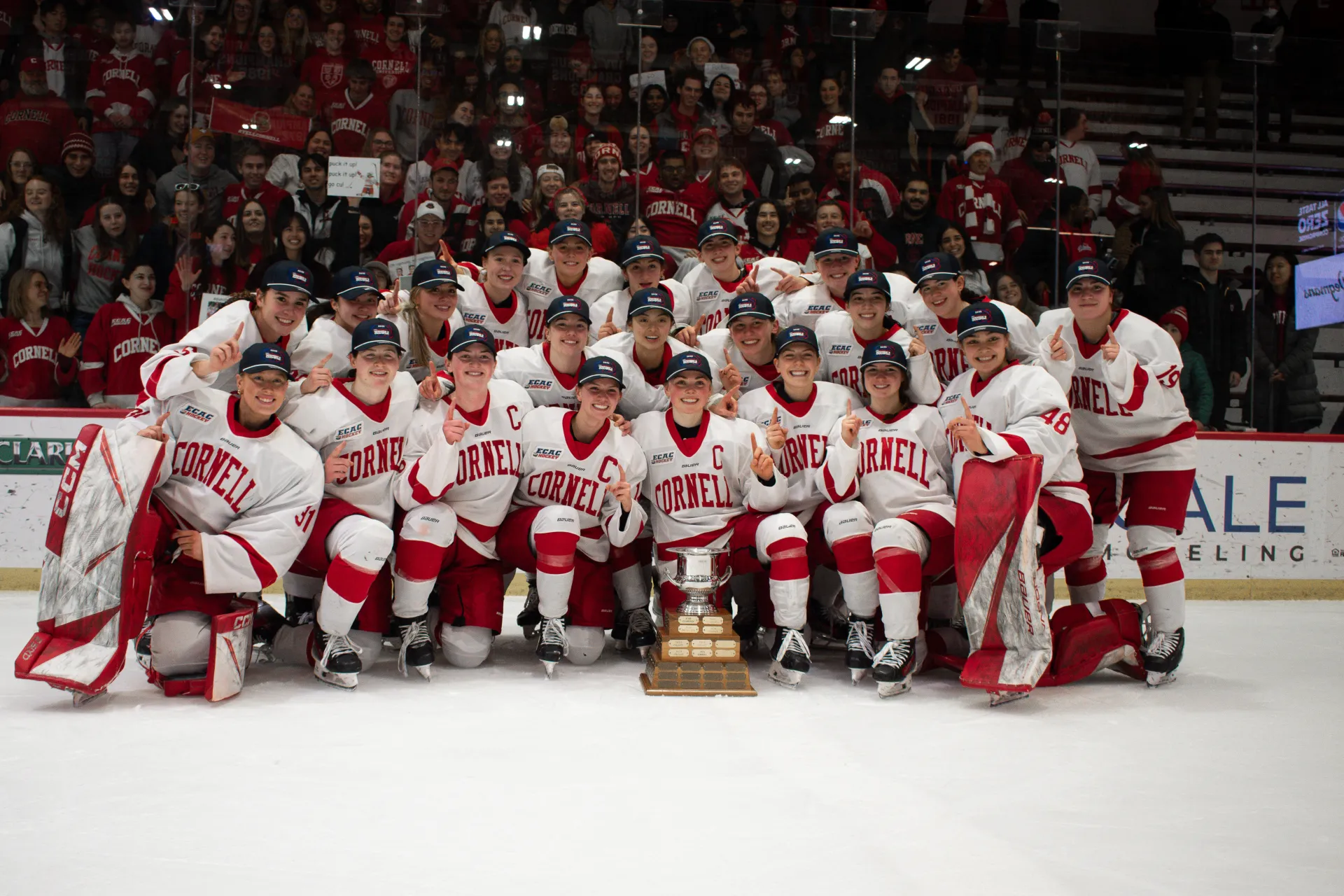 Women's hockey team in uniform and ECAC championship hats gathered for large group photo on ice with trophy